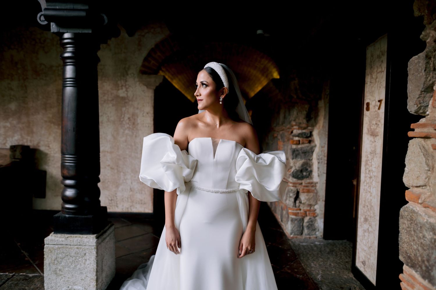 Elegant bride in an off-shoulder white wedding gown with dramatic puff sleeves poses in a stone archway.