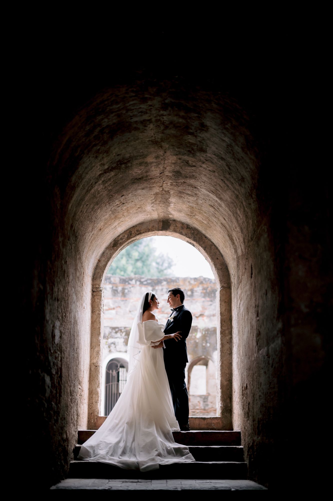 Silhouetted wedding couple standing in a dramatic stone archway with bright natural backlighting.