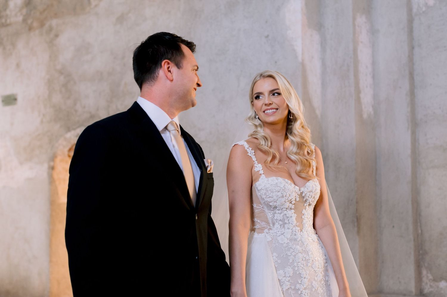 Joyful wedding portrait of couple in formal attire against weathered stone wall.