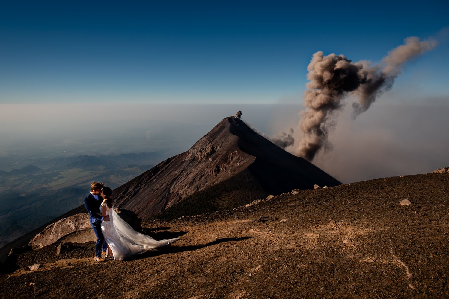 A couple poses on a rocky slope while the active Fuego volcano erupts with smoke plumes behind them in Guatemala.