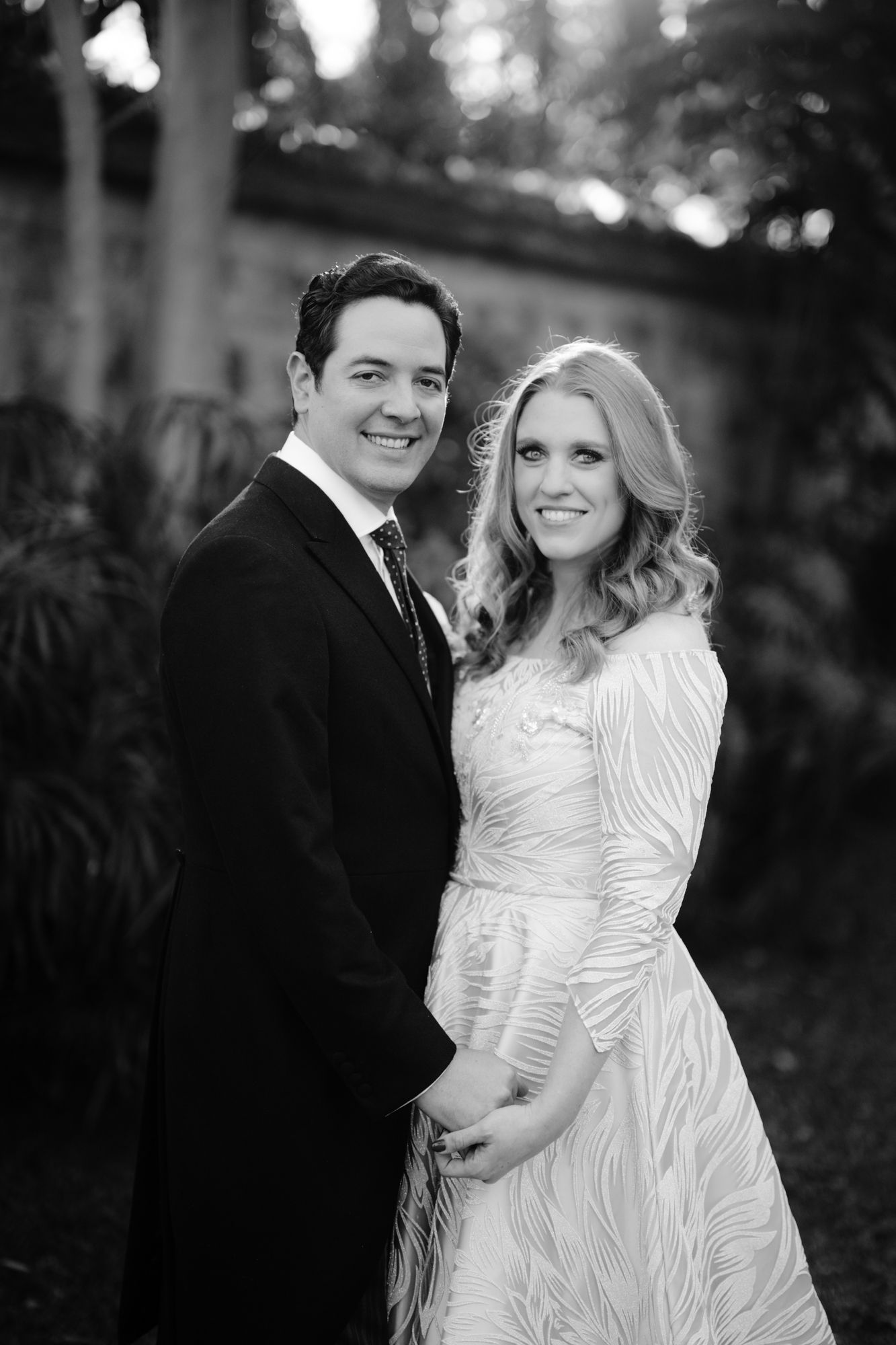 A bride in a white patterned dress poses with her groom in a black tuxedo outdoors in a garden setting.