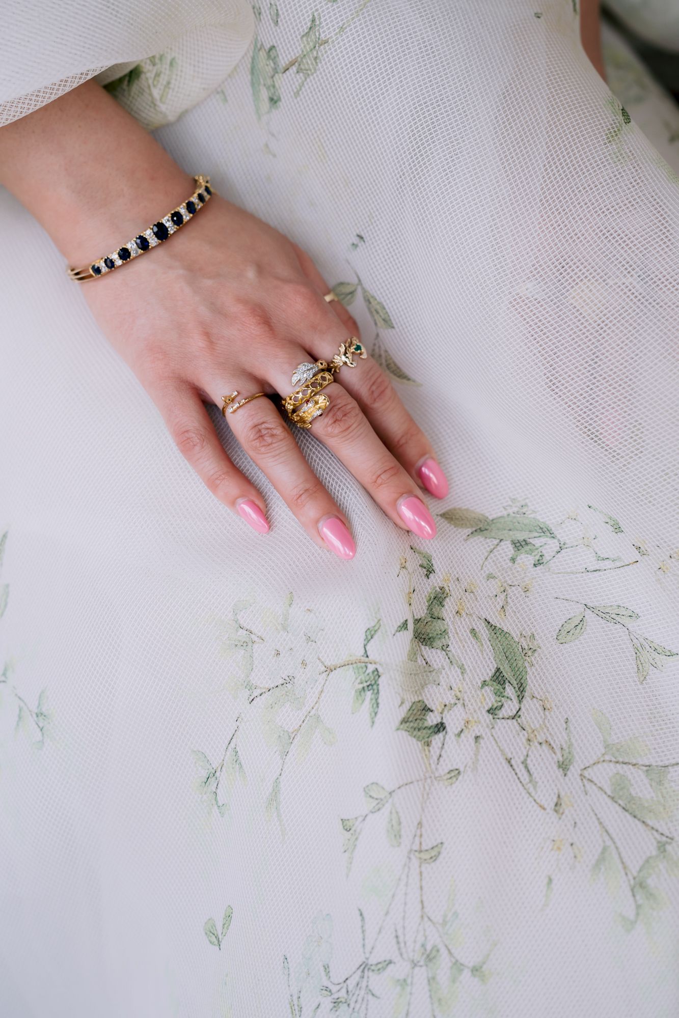 Close-up of pink manicured hand wearing gold jewelry and rings against white fabric with delicate floral pattern.