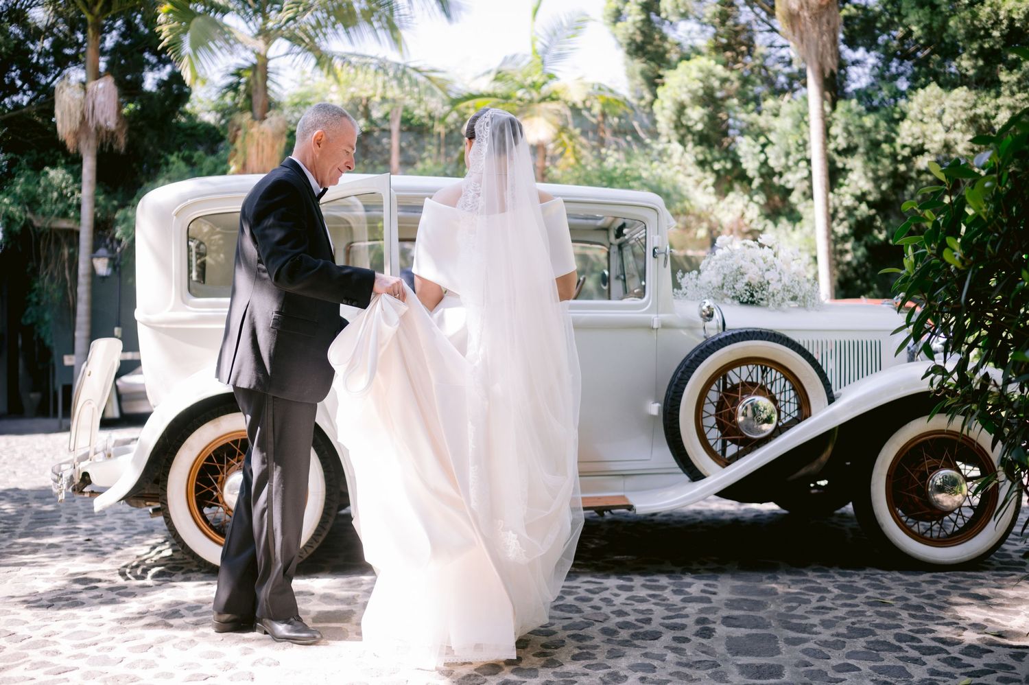 Elegant wedding moment beside a pristine vintage white automobile under palm trees.