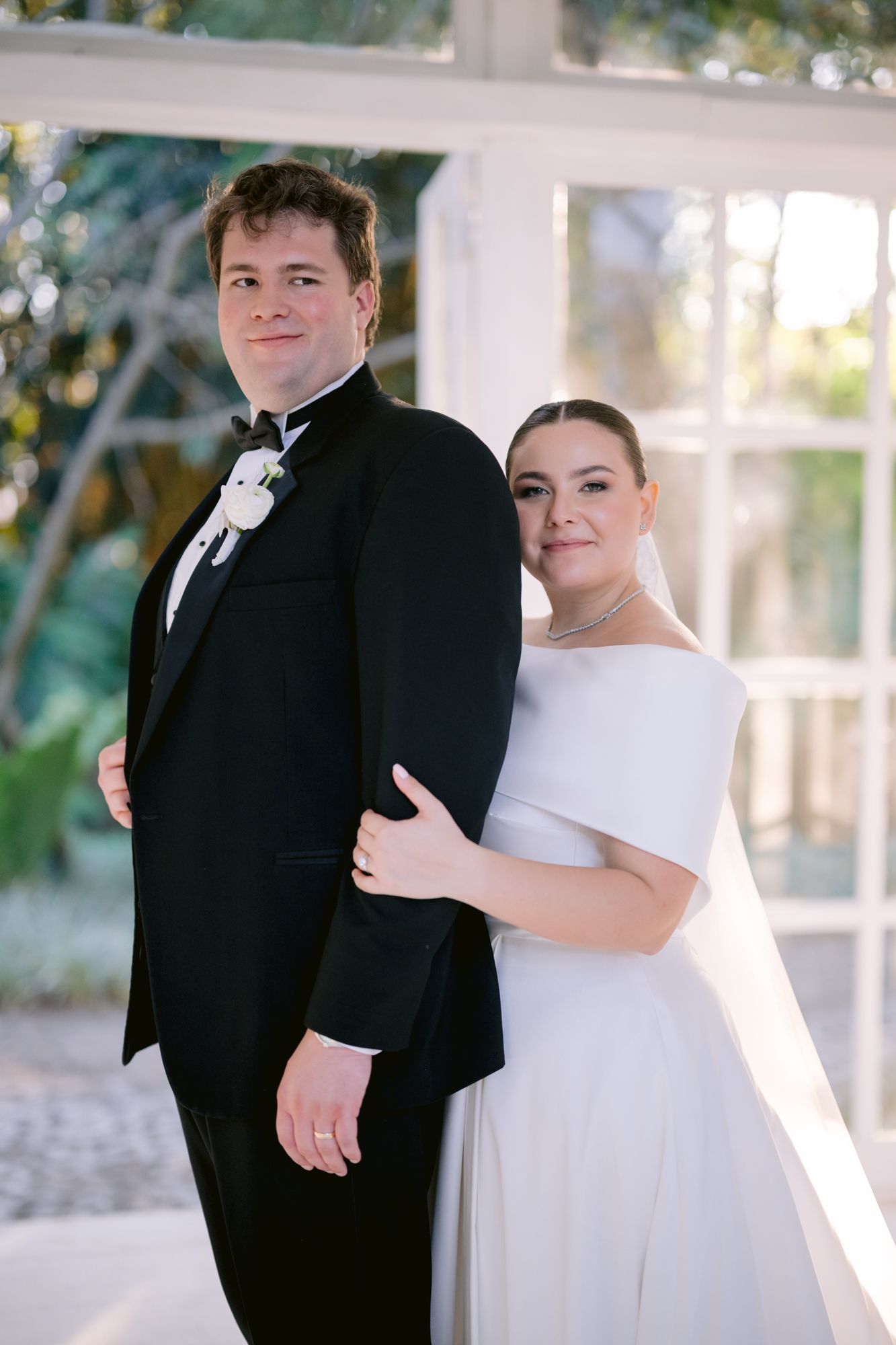 Elegant bride in a simple white dress poses with groom in classic black tuxedo against bright windows.