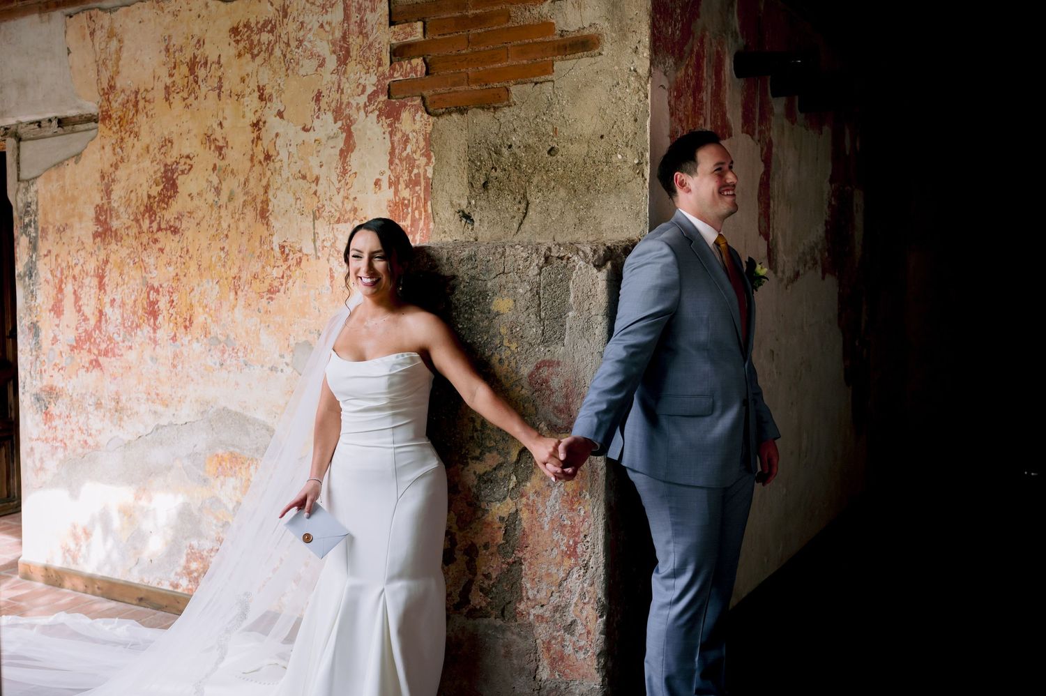 Romantic wedding photo of couple holding hands against a rustic textured wall in an artistic composition.