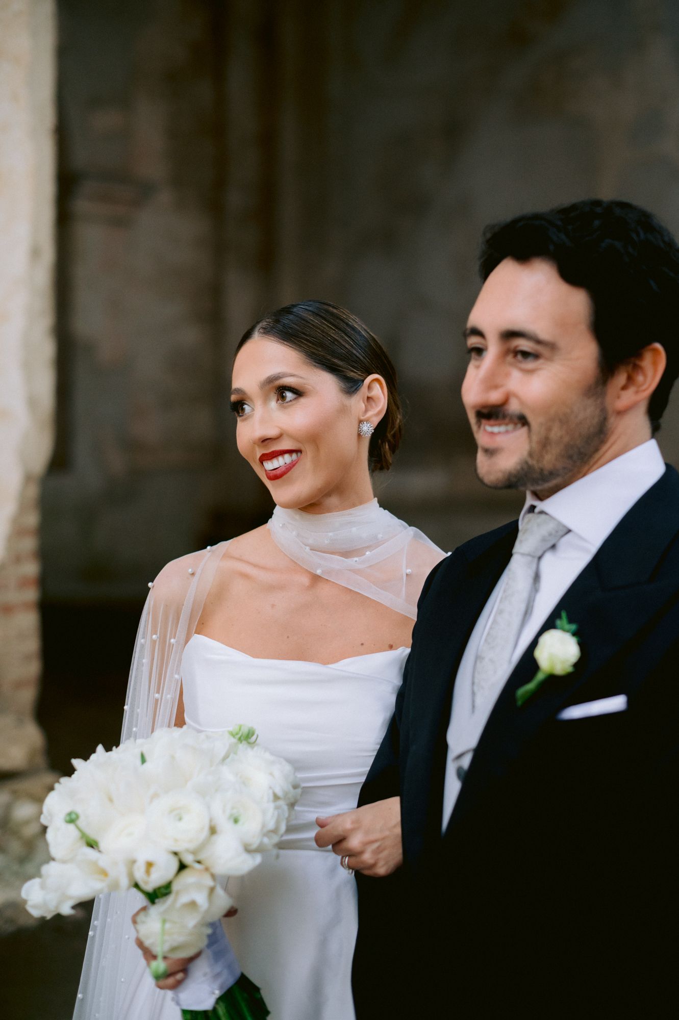 Elegant bride in high-neck wedding gown holds white bouquet while standing with groom.