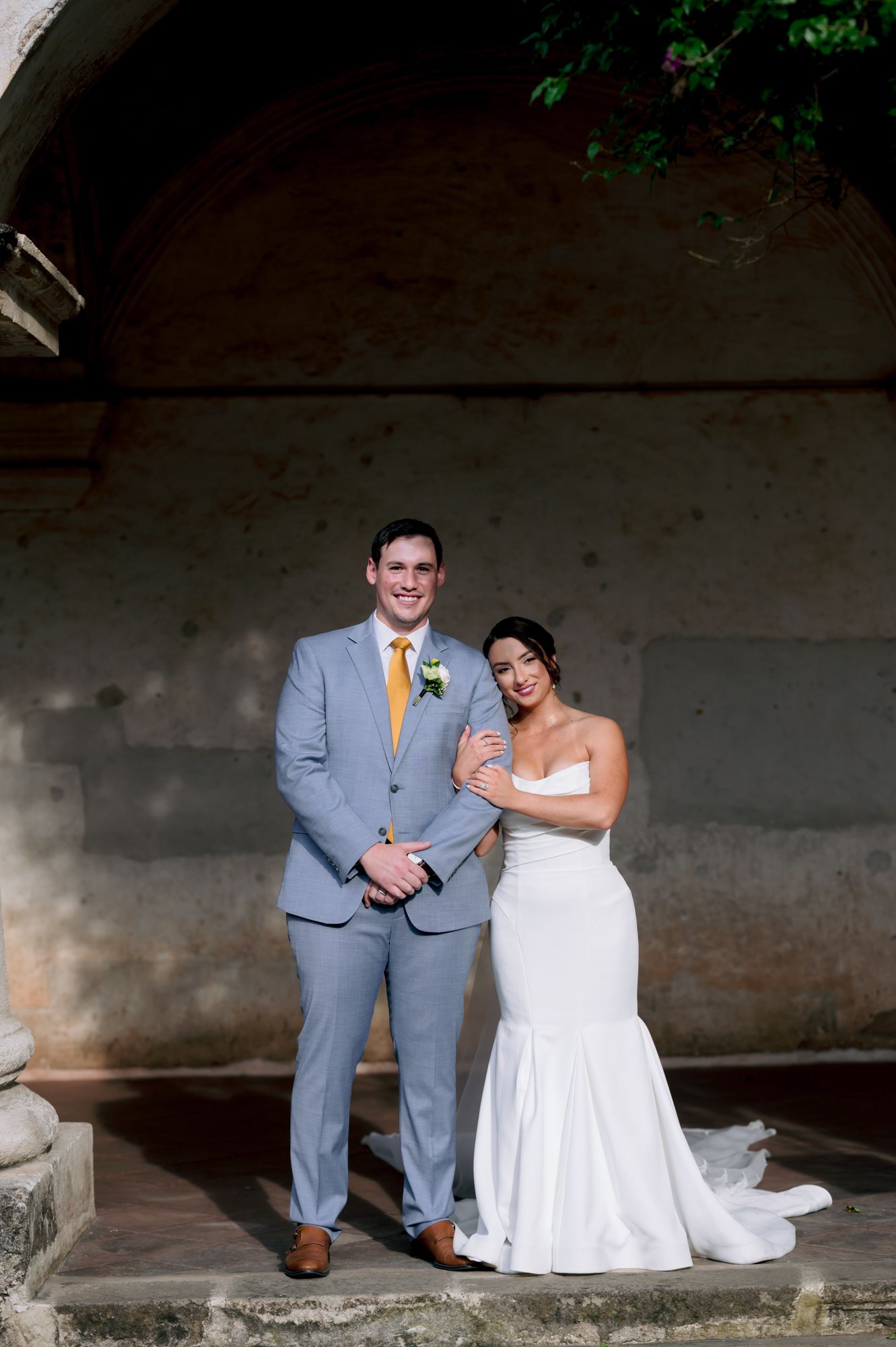 Wedding couple poses under stone archway in classic formal attire.