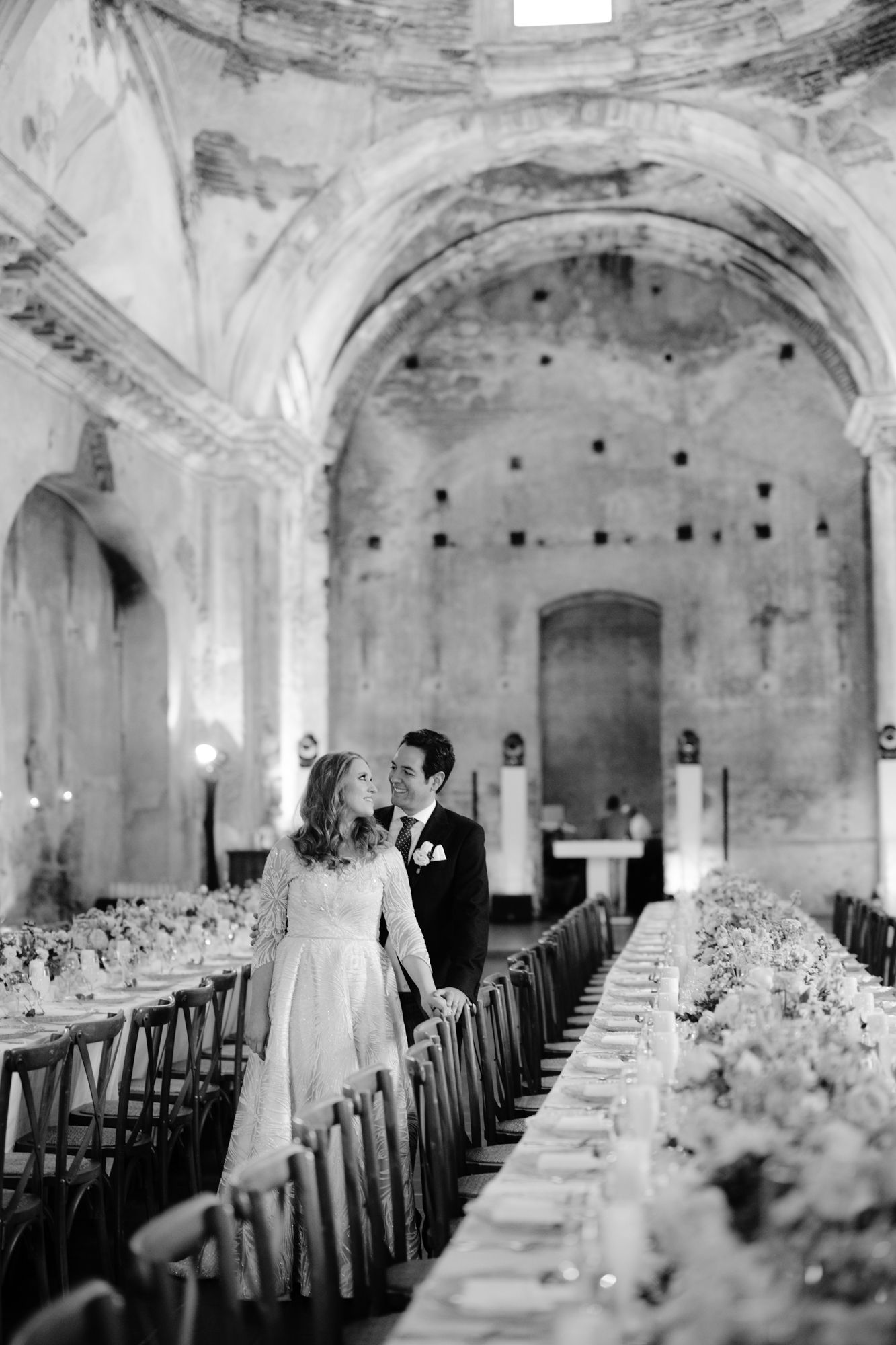 A black and white photo of a couple among elegant table settings in a historic venue.