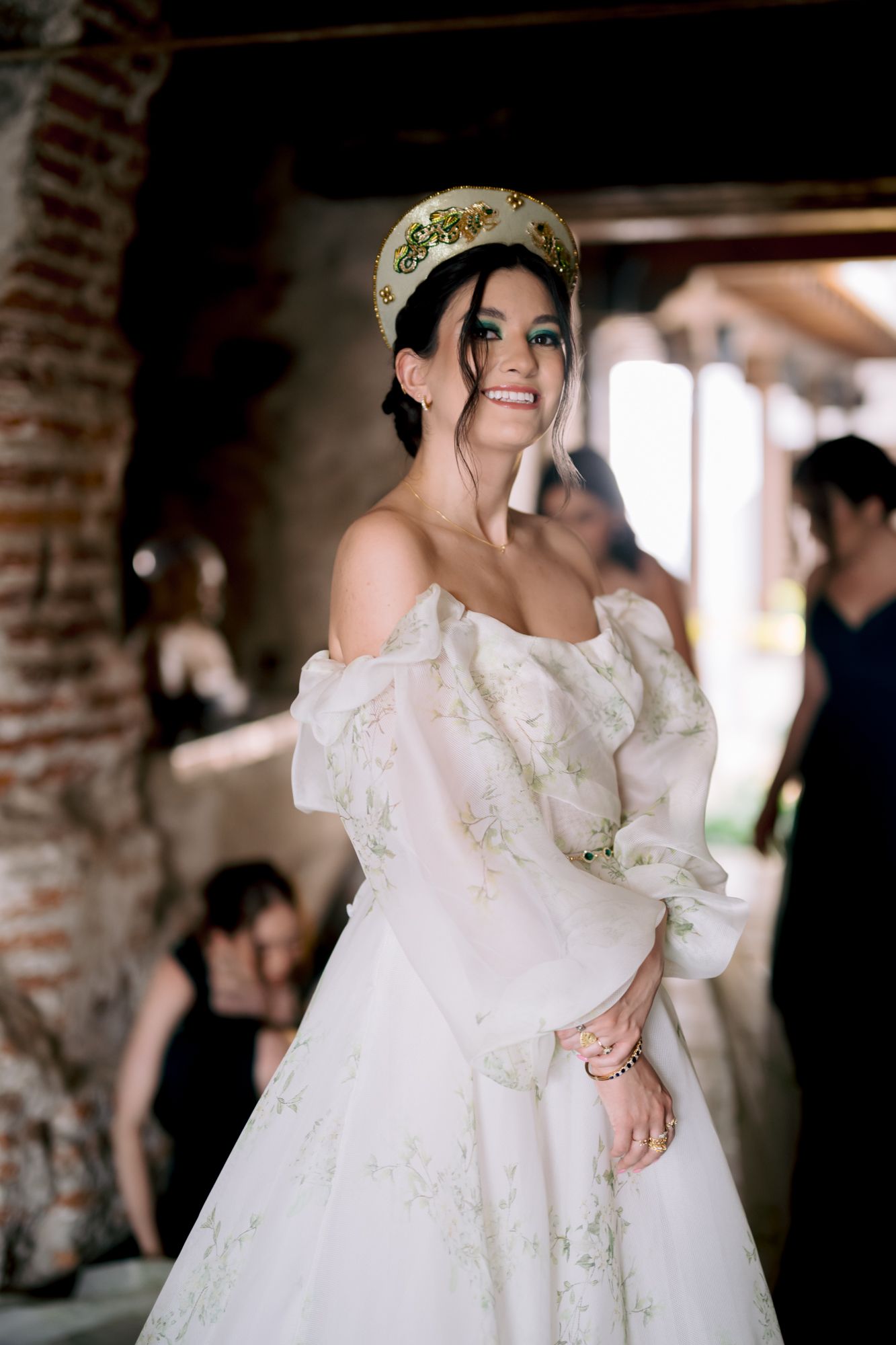 A person wearing an ornate crown and off-shoulder wedding gown smiles radiantly in a rustic venue.