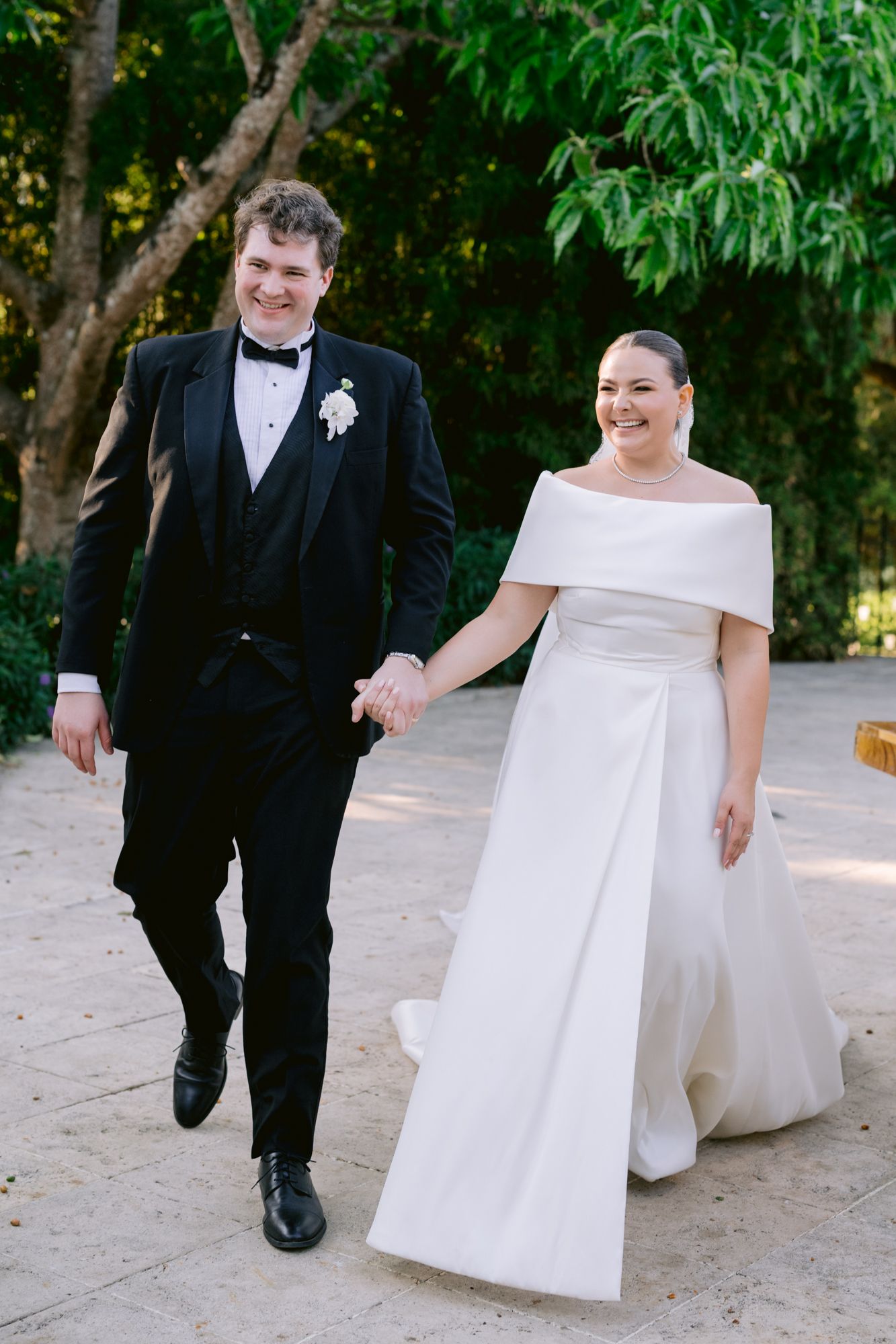 Bride in off-shoulder gown and groom in tuxedo walking hand-in-hand outdoors.