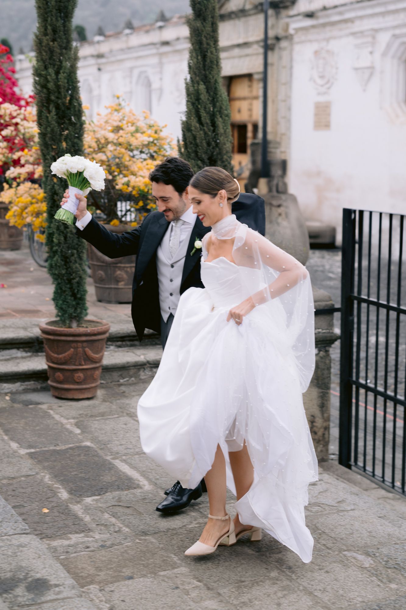 Newlyweds walking together outdoors with bride holding white bouquet and groom assisting with dress.