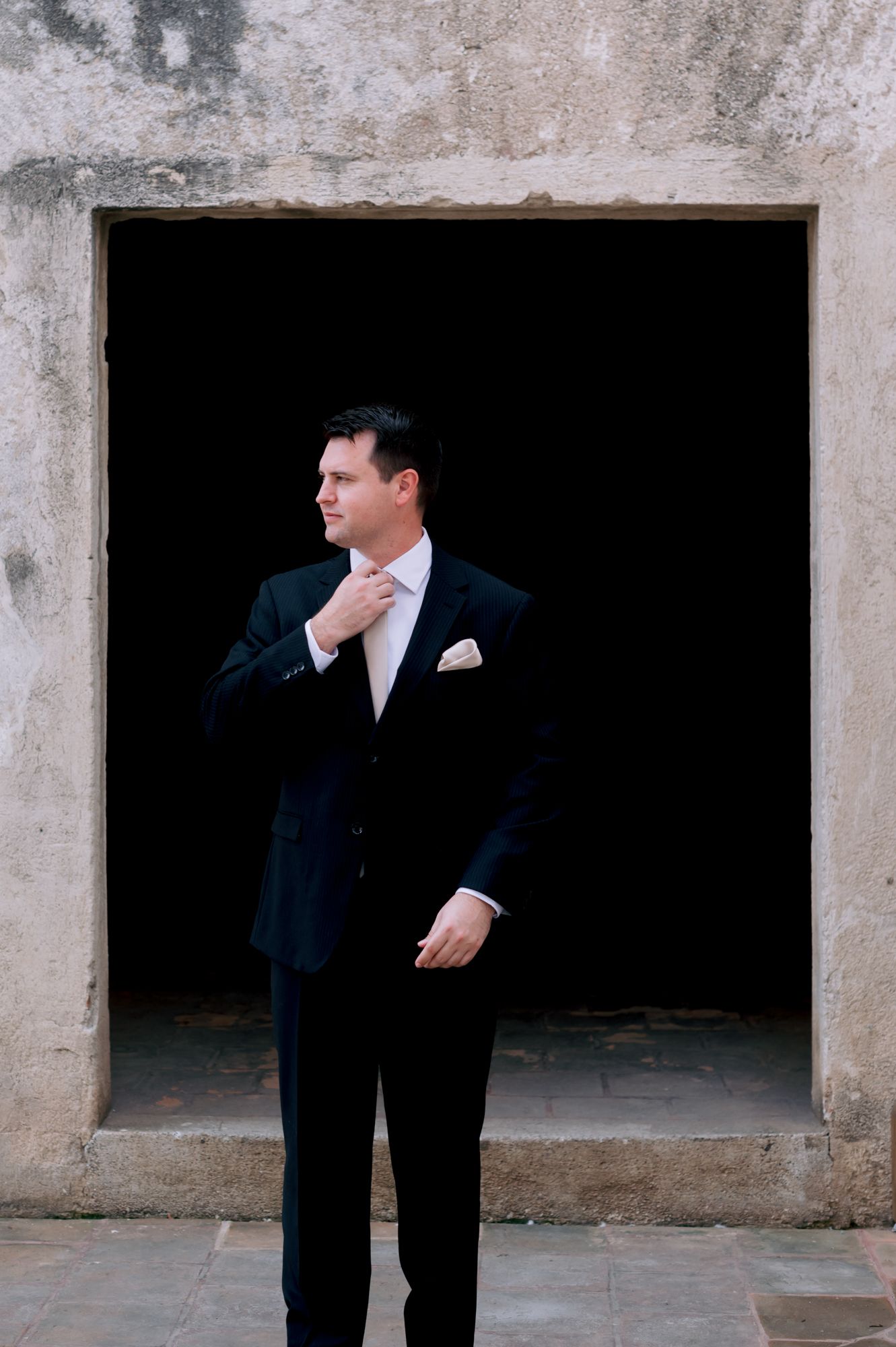 A groom adjusting his tie while standing in a dramatic concrete doorway.