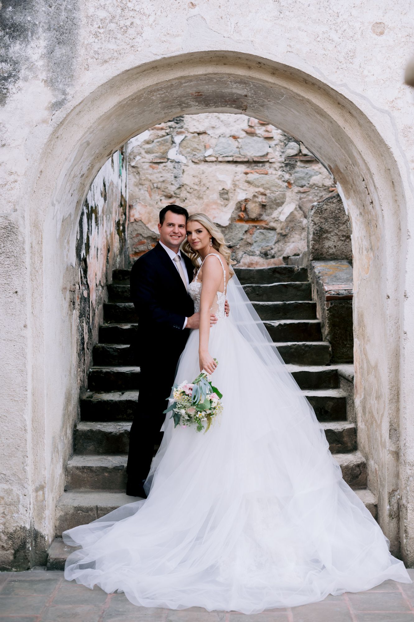Wedding couple poses under stone archway with bride in white gown holding blue bouquet.