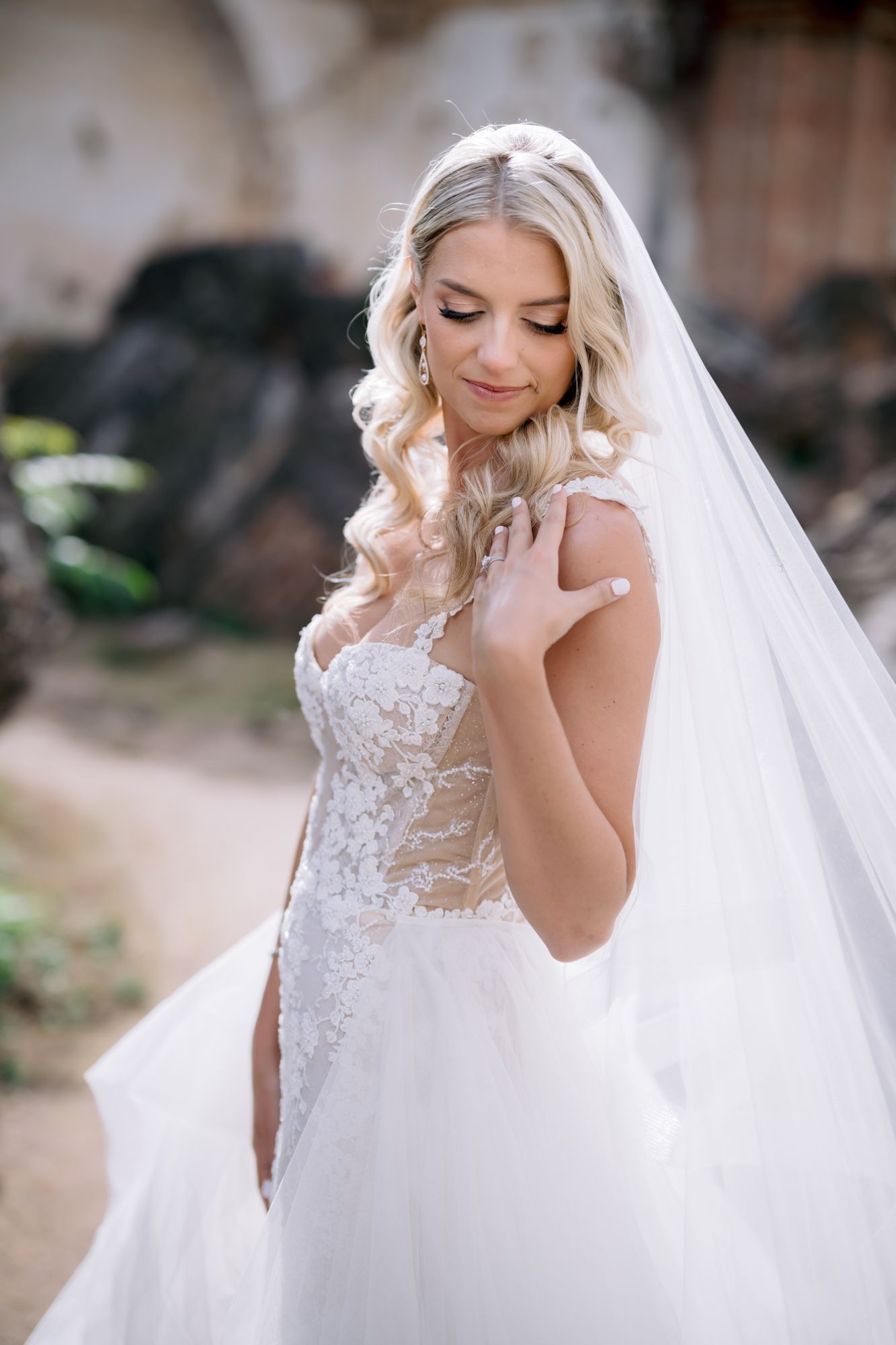A bride in a detailed white lace gown with long veil poses gracefully at historic venue.