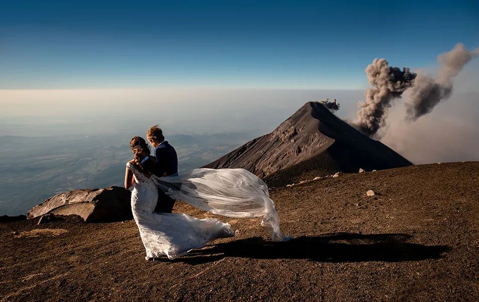 A couple embraces on a volcanic peak with dramatic smoke plumes rising from an active crater in the background.