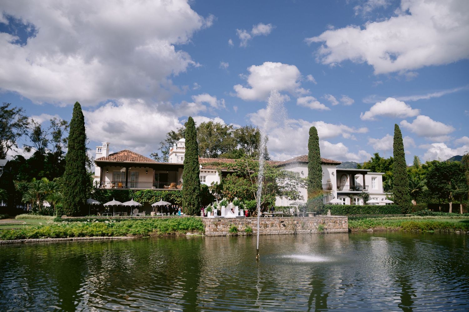 Elegant waterfront venue with cypress trees and white architecture reflected in a tranquil lake on a sunny day.
