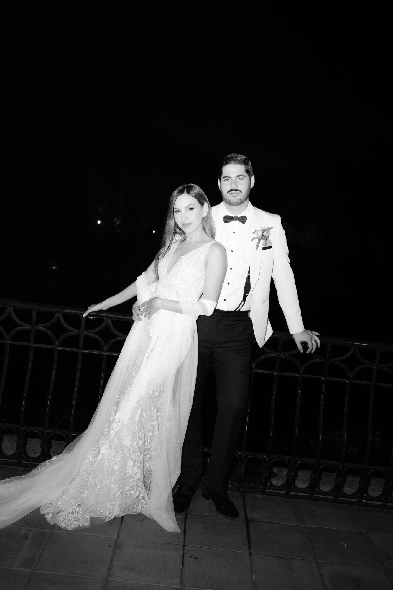 A black and white photo of a bride in a lace gown and groom in white tuxedo jacket posing together at night.