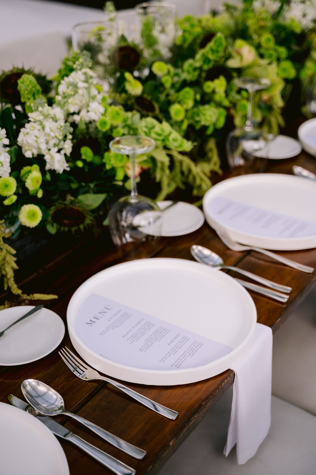 Elegant table setting with white plates, cutlery and fresh green floral centerpieces on dark wooden tables.