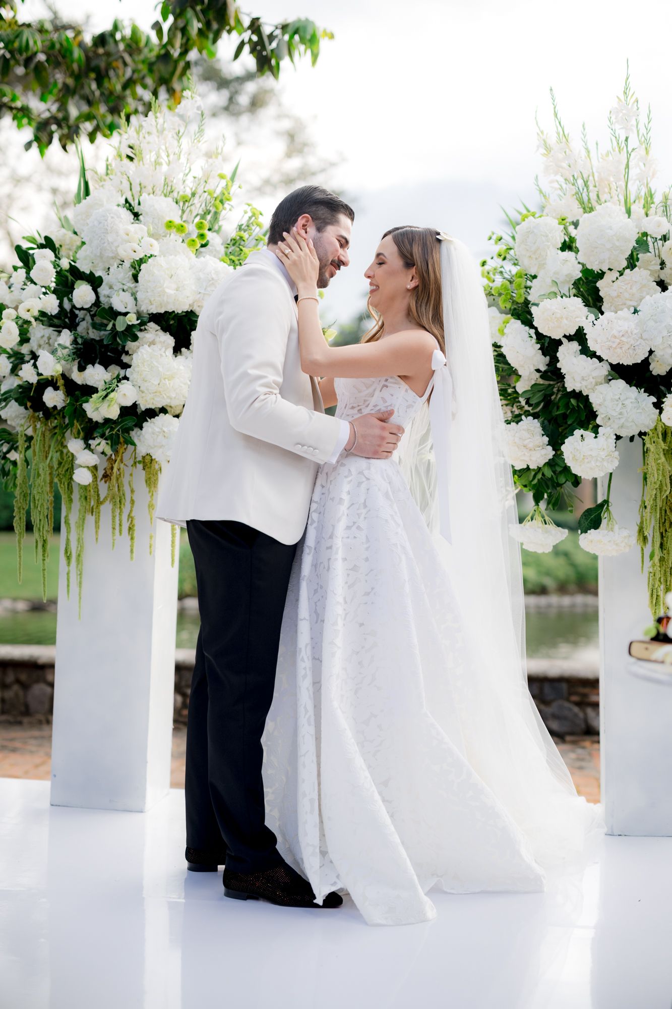 Elegant wedding ceremony setup with white floral arch and couple embracing on white platform.
