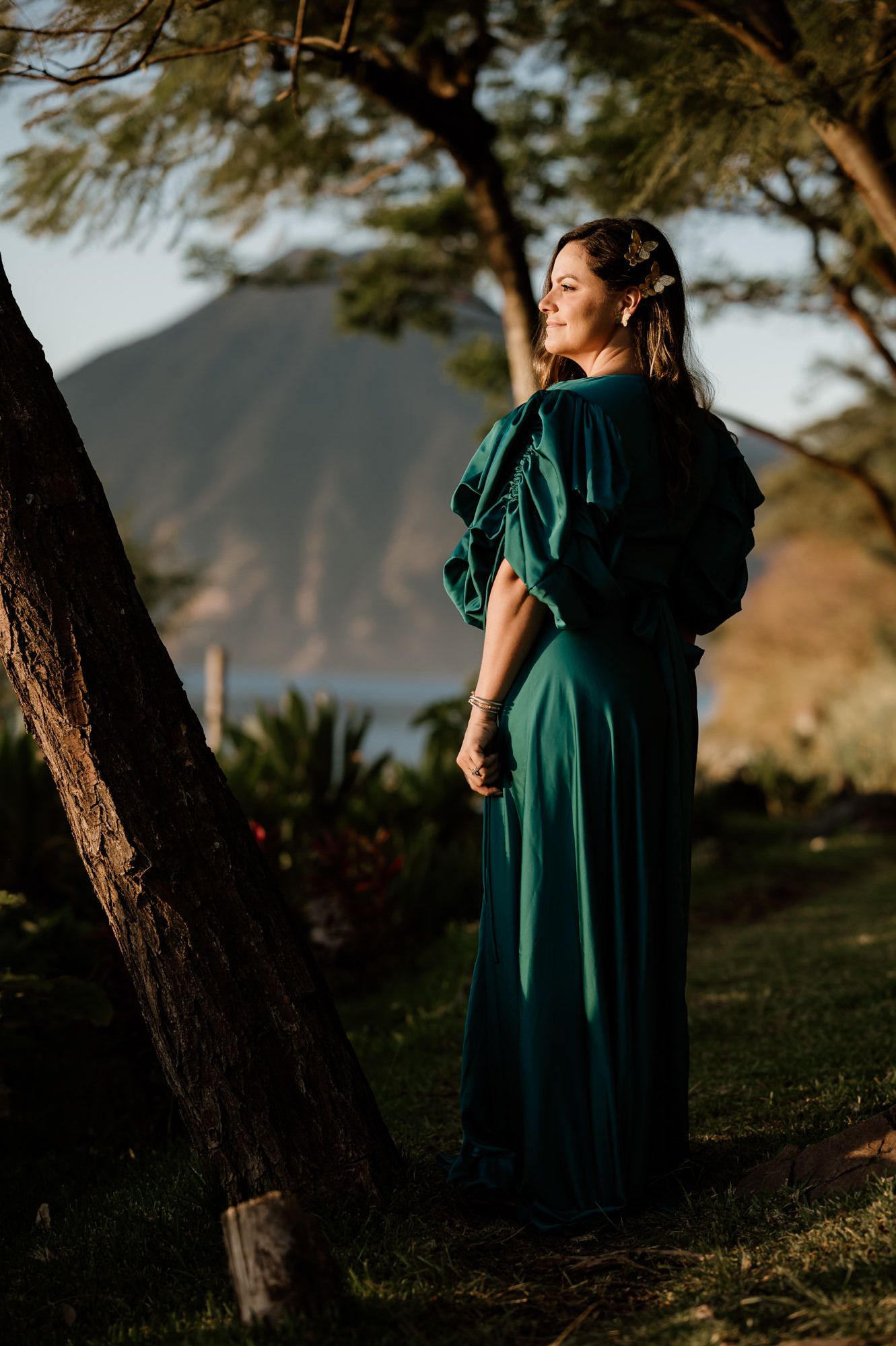 A woman in an emerald green dress poses against a tree during golden hour in a scenic outdoor setting.