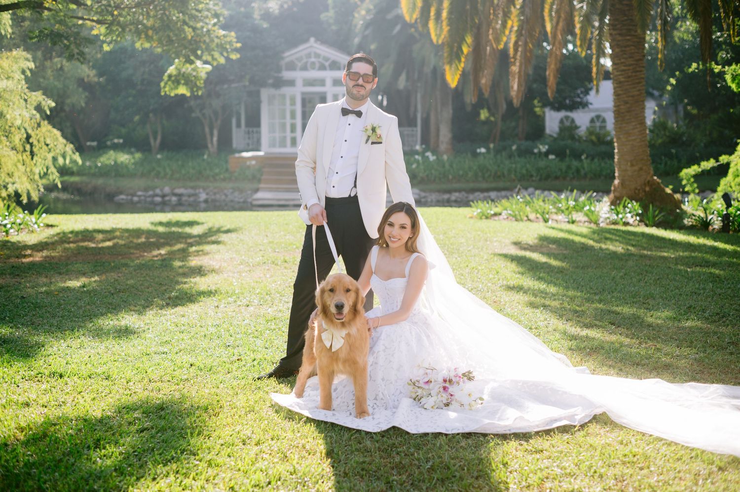 A couple and their golden retriever dog pose for outdoor wedding photos in a sunlit garden setting.