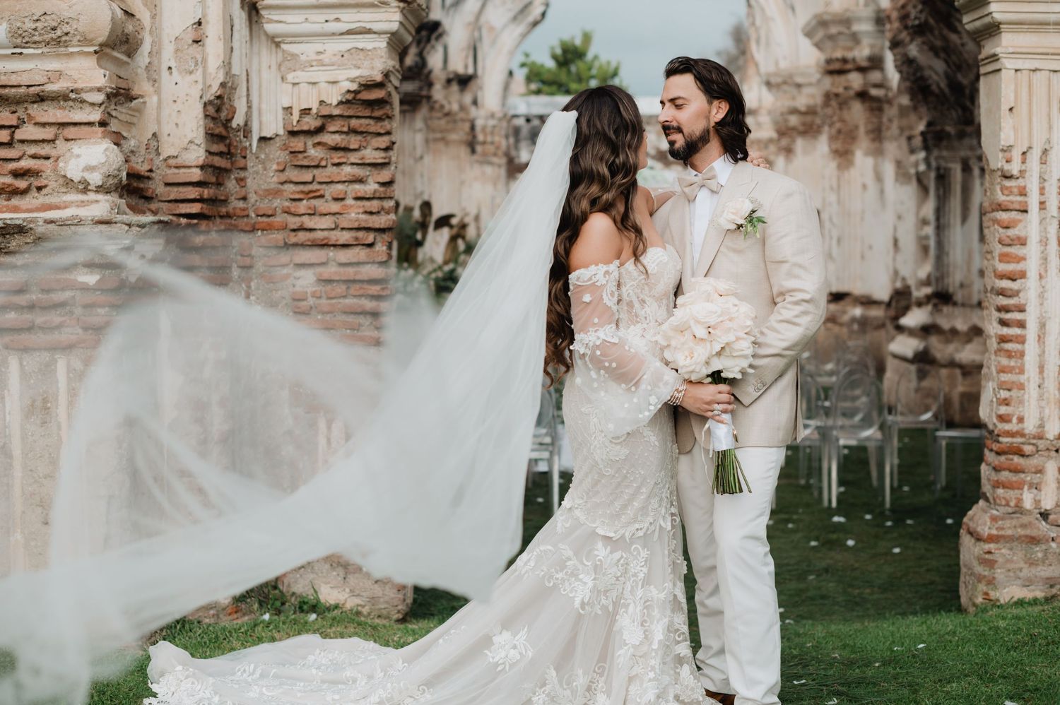 Flowing white wedding dress and veil create movement against rustic stone ruins backdrop.