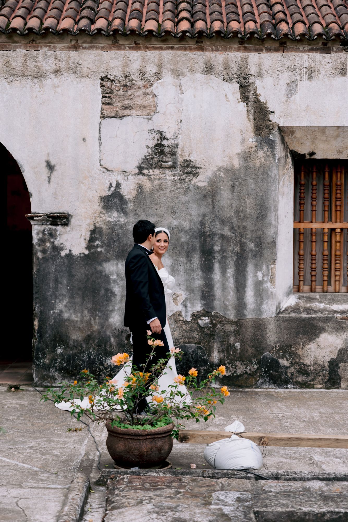 Newlyweds pose against weathered colonial wall beside potted flowers in rustic venue.