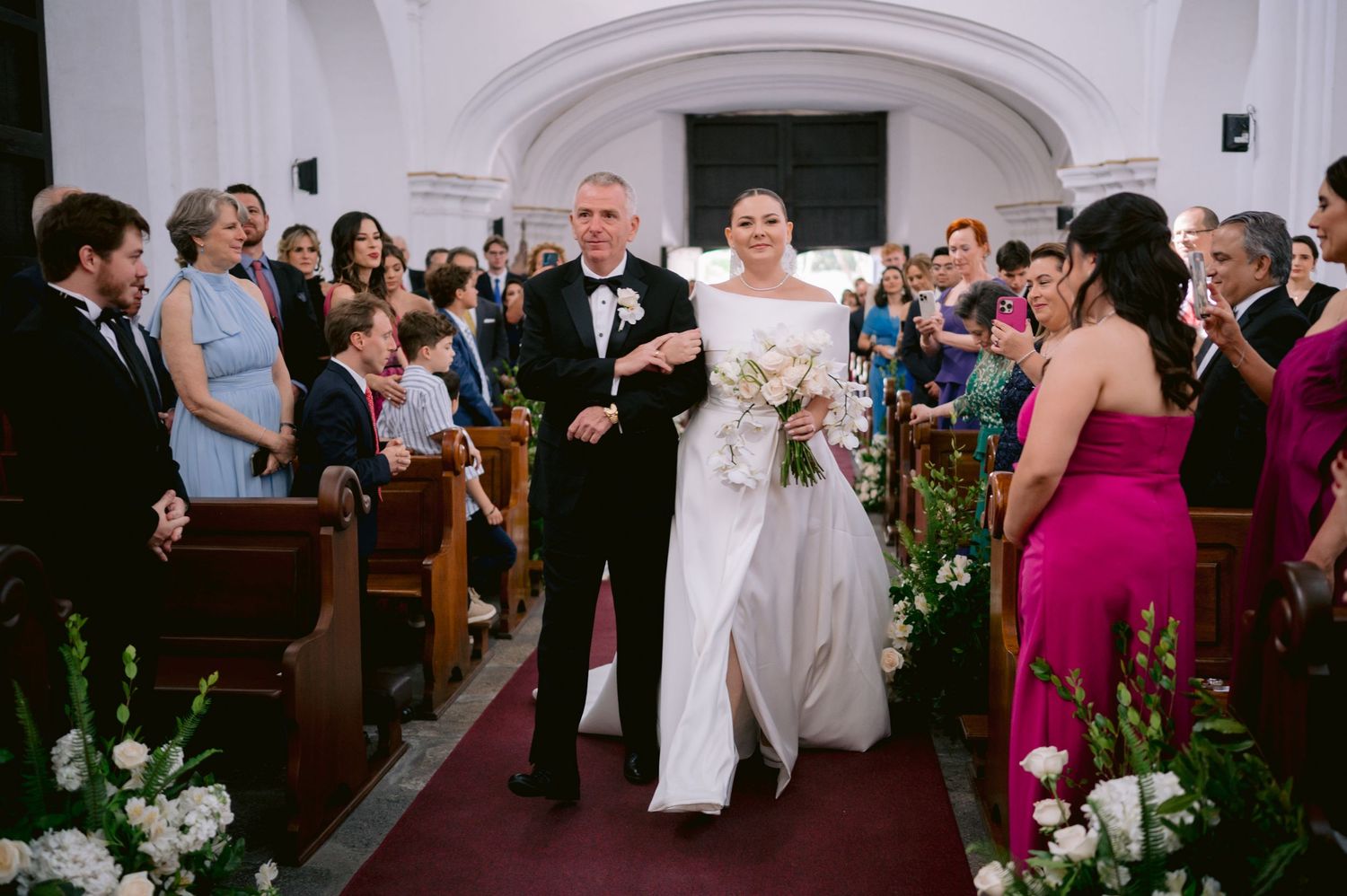 Wedding processional in a traditional church with floral decorations lining the aisle.