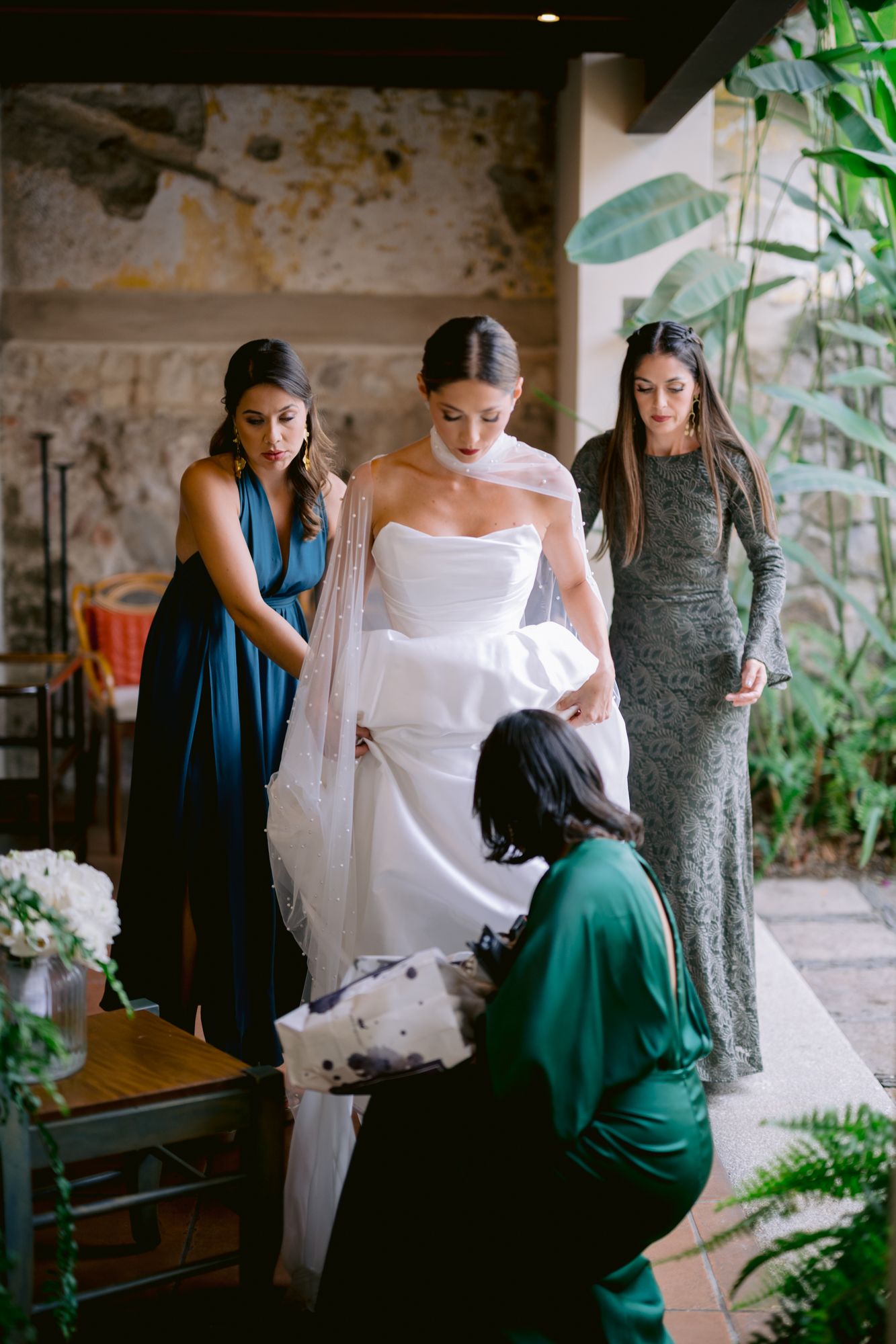 A wedding dress fitting session with bridesmaids in blue and grey dresses assisting the bride in a garden patio setting.