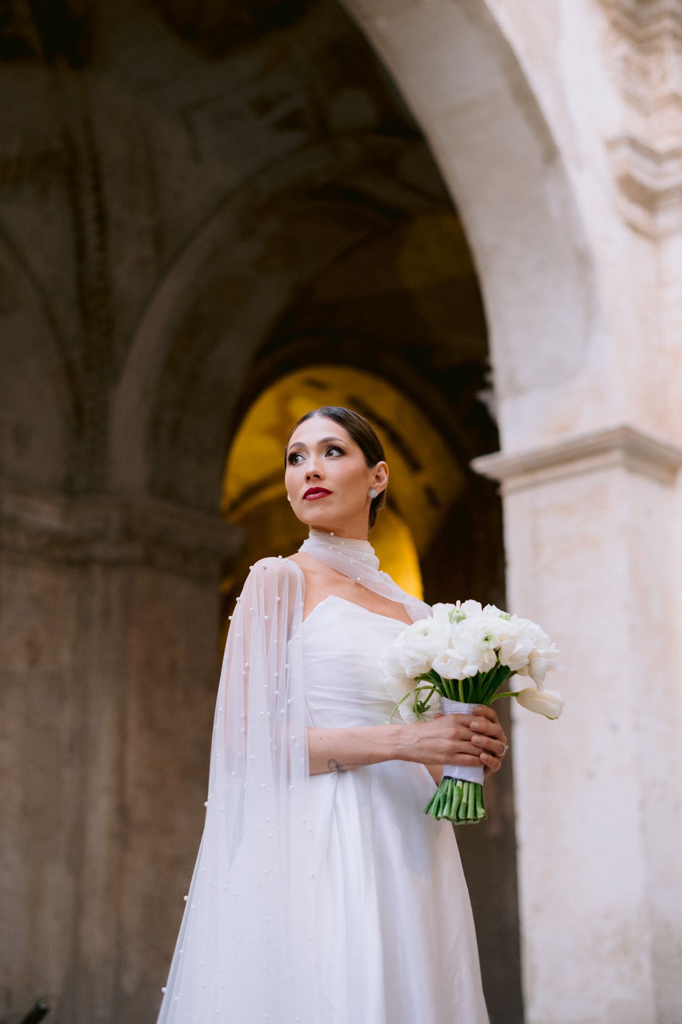 Bride holding white bouquet poses in cathedral with dramatic arched columns and golden lighting.