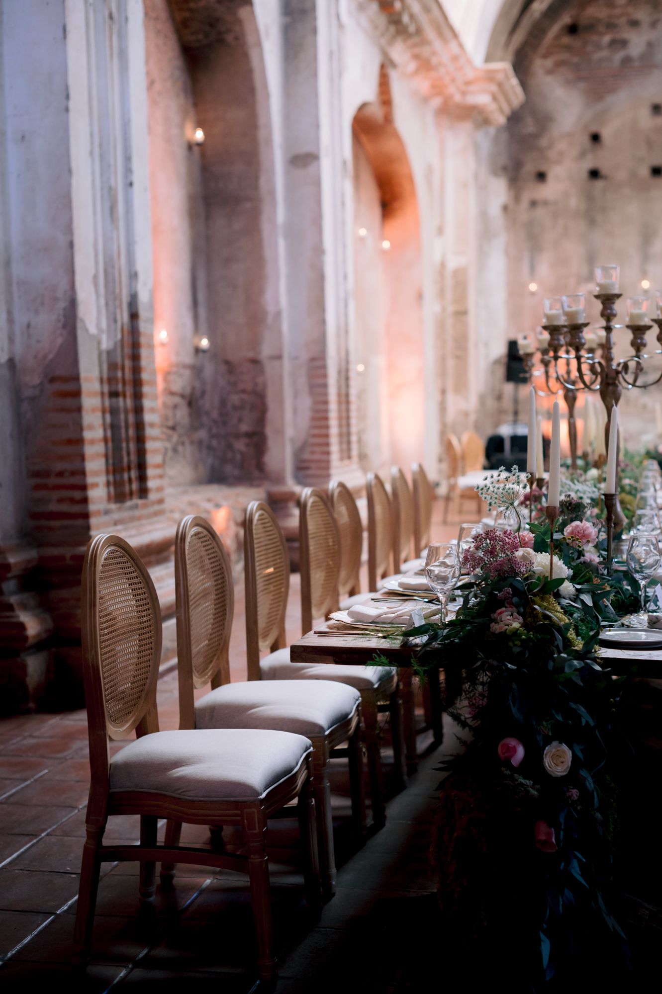 Long dining table with cane back chairs and floral arrangements set in historic church interior.