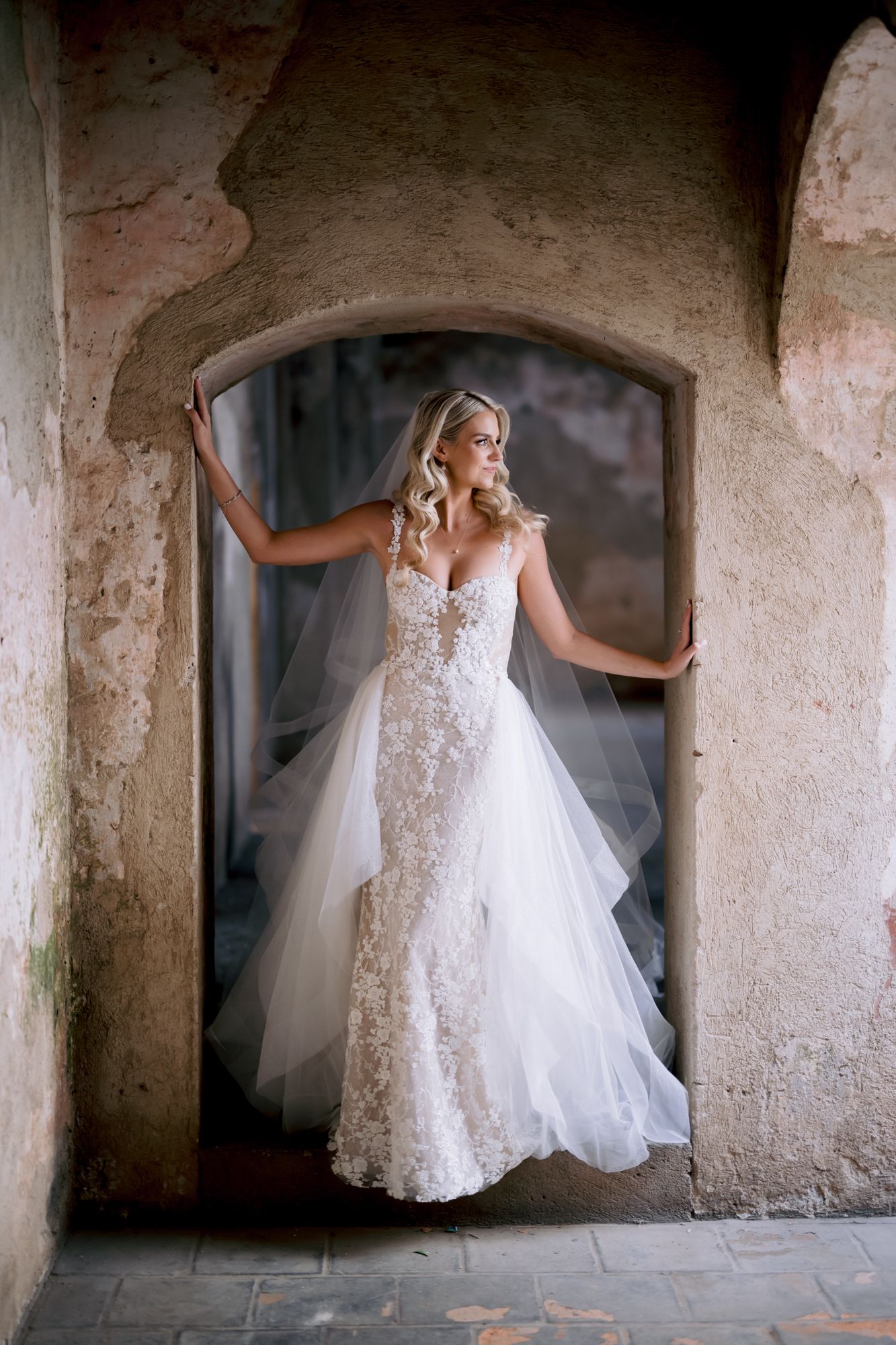 A bride in a fitted lace gown poses gracefully in an ancient stone archway.
