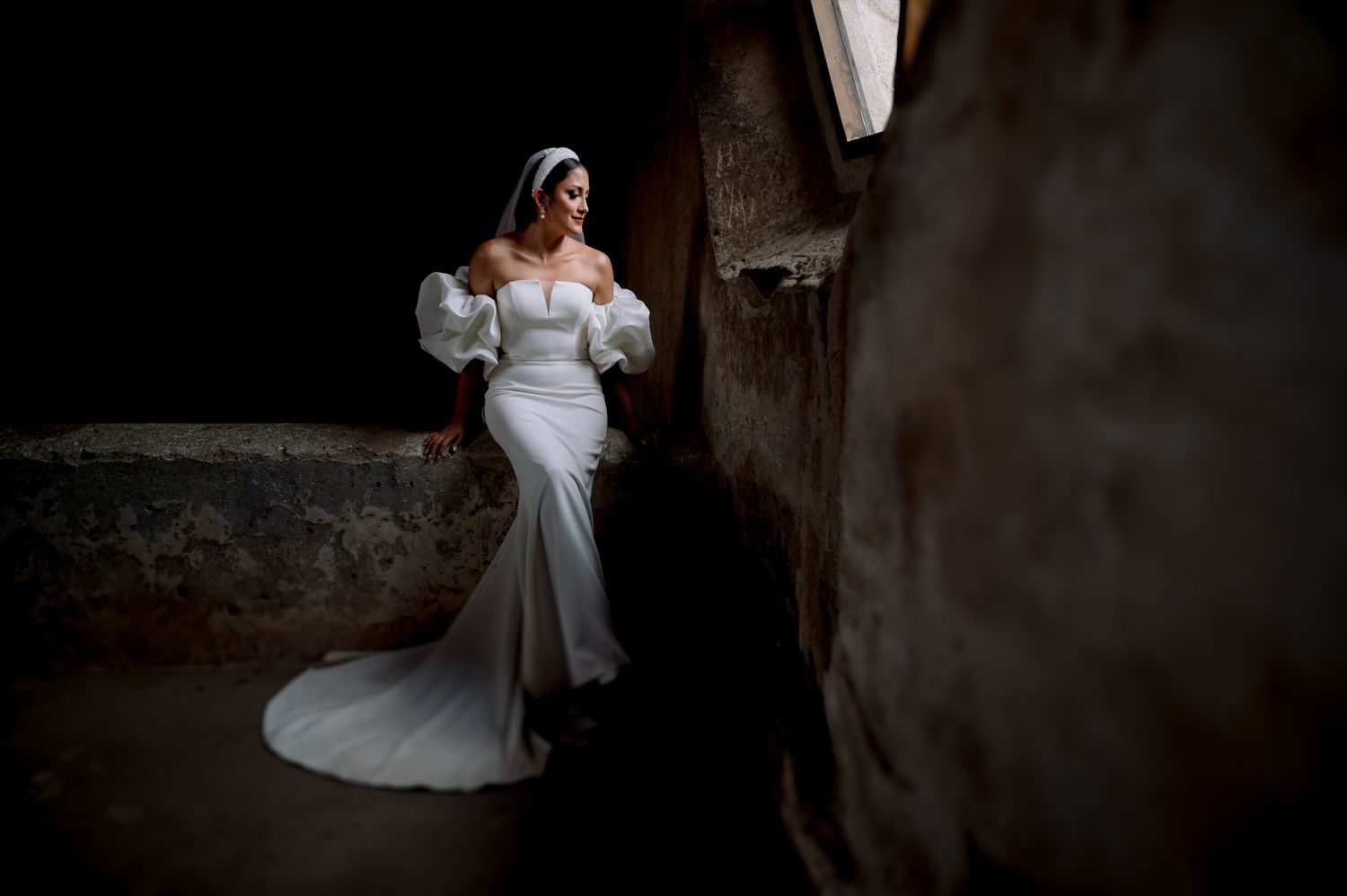 Dramatic lighting captures bride in form-fitting gown with statement sleeves against textured wall.