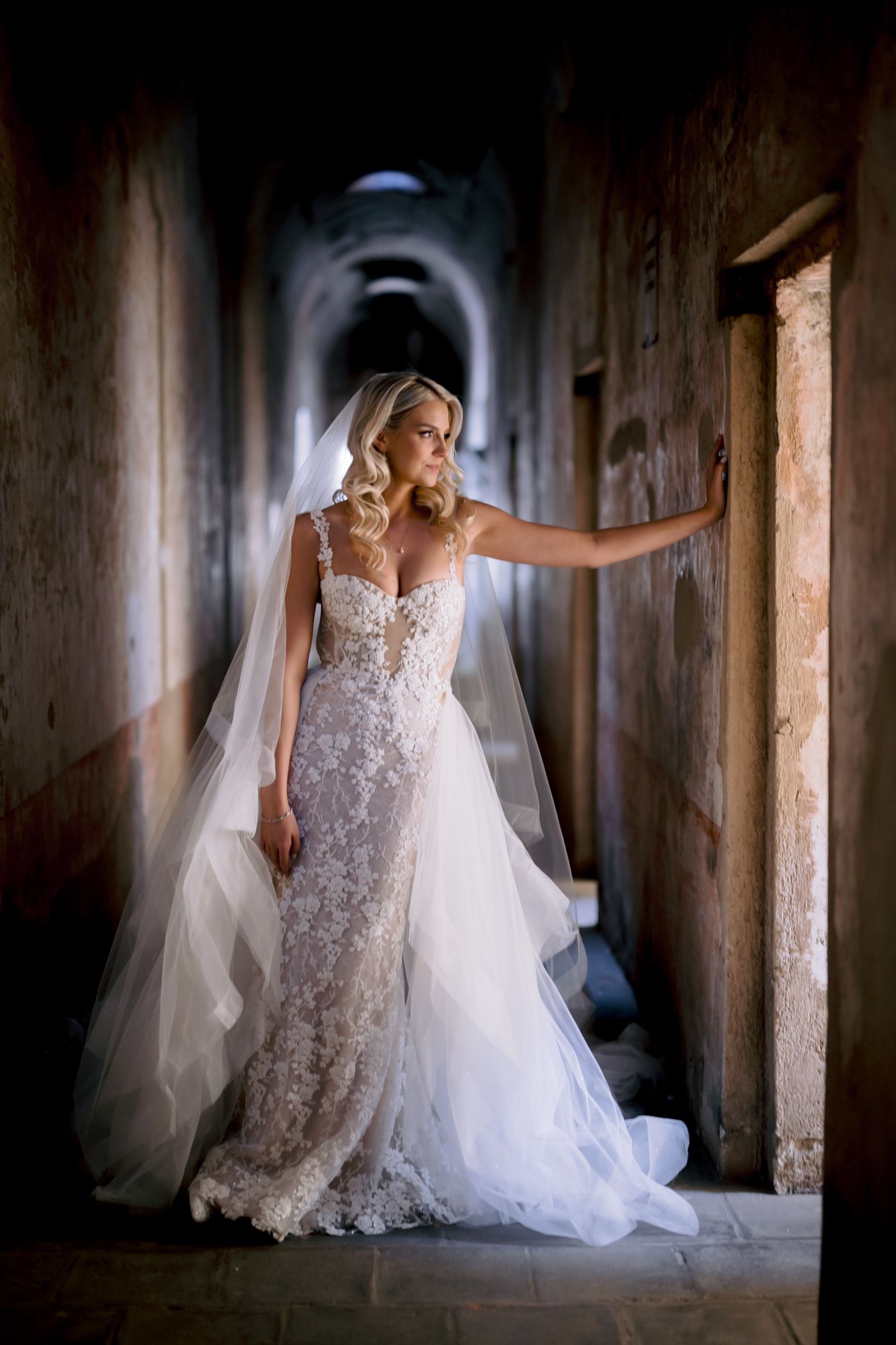 Bride in lace gown poses in dramatic corridor with natural light streaming from above.