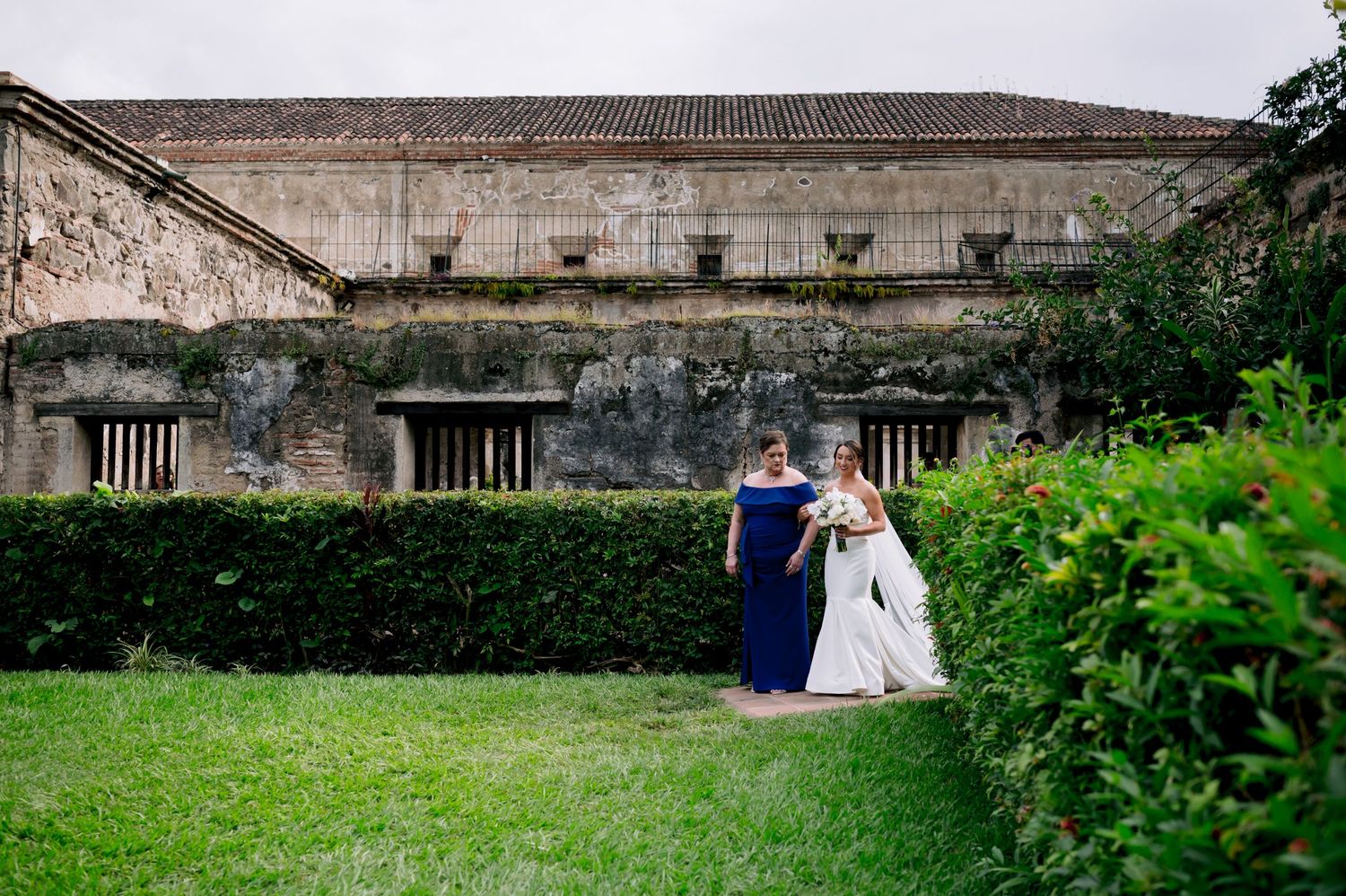 A couple poses for photos in front of a weathered stone building with lush greenery.