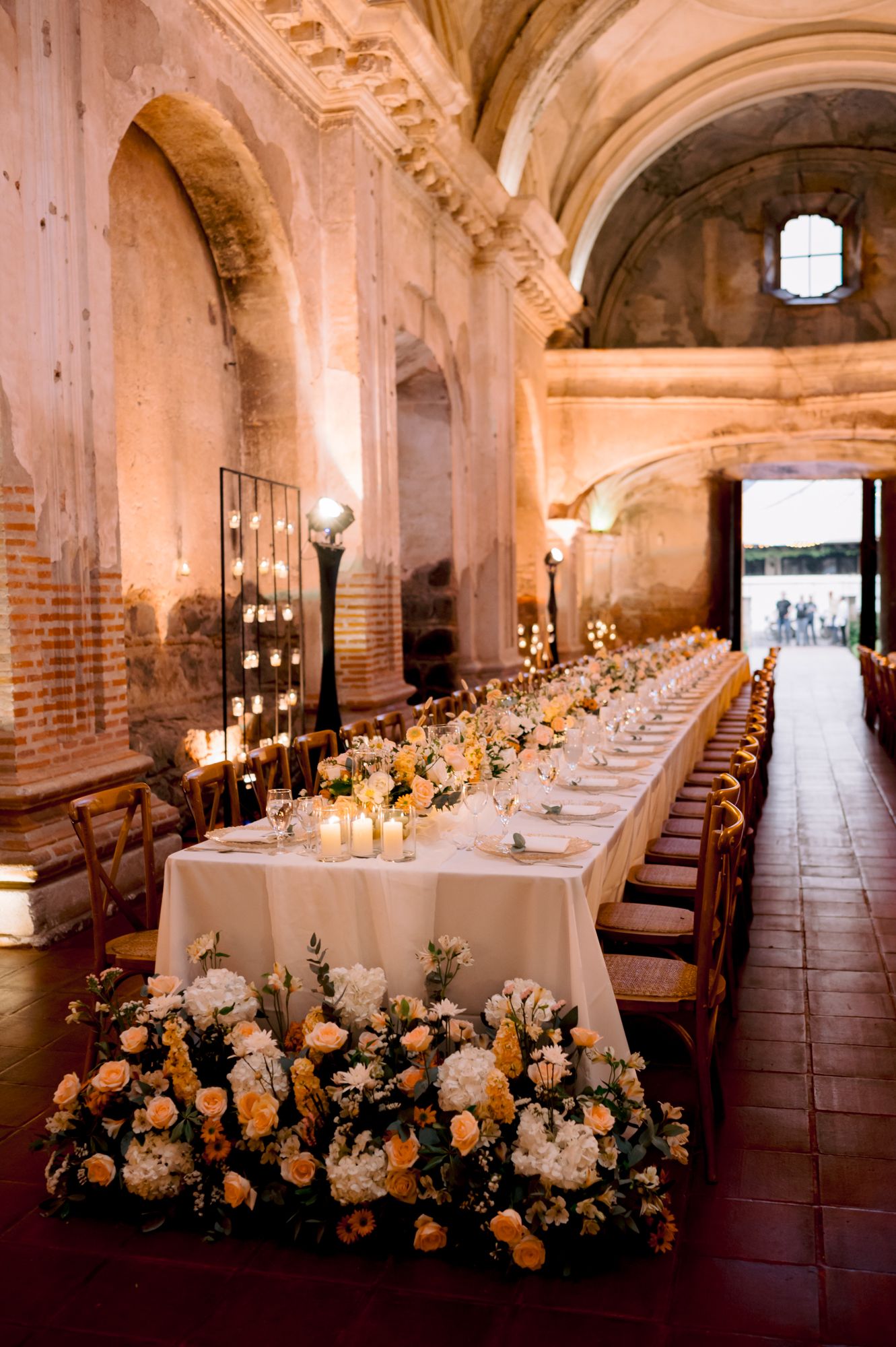 Long banquet table with white linens and floral centerpieces in historic stone chapel setting.