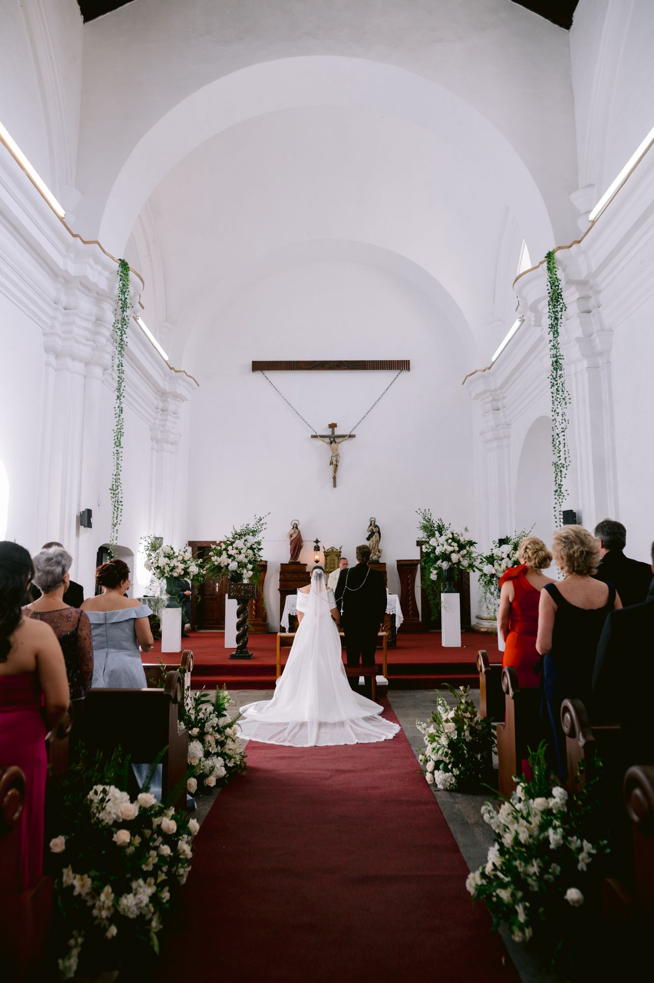 An elegant church wedding ceremony with white floral arrangements lining the aisle and crucifix at the altar.