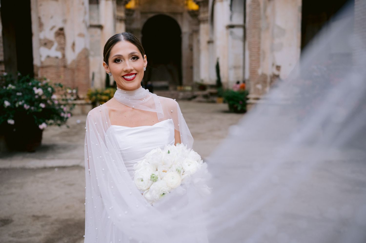 Bride with flowing cape and white bouquet smiles in historic church setting.