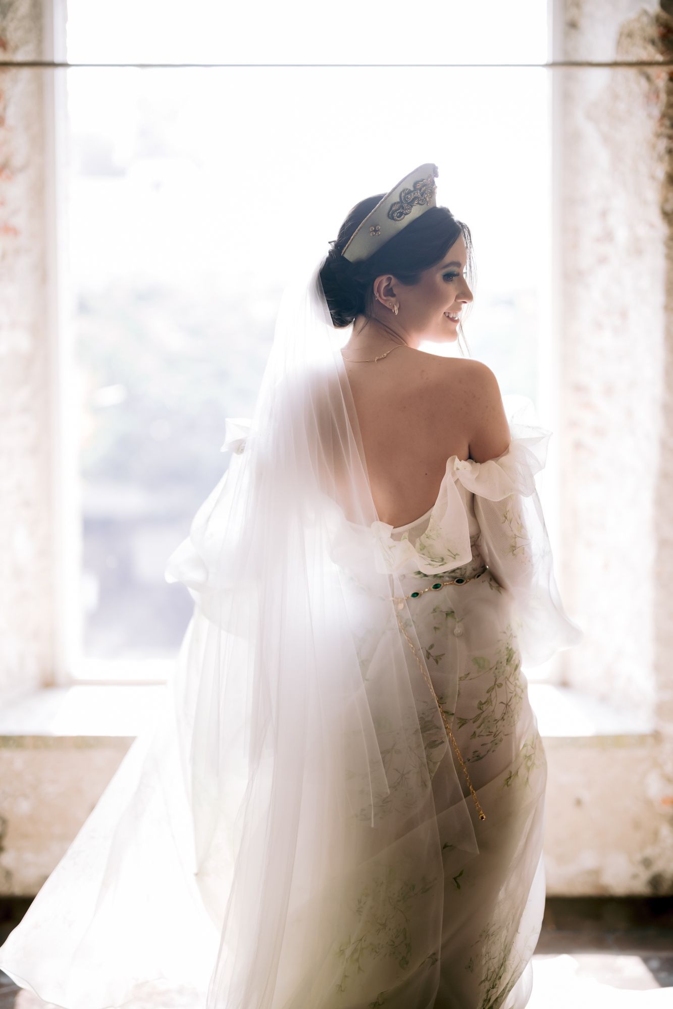 Romantic back-facing bridal portrait with flowing veil against bright window light.