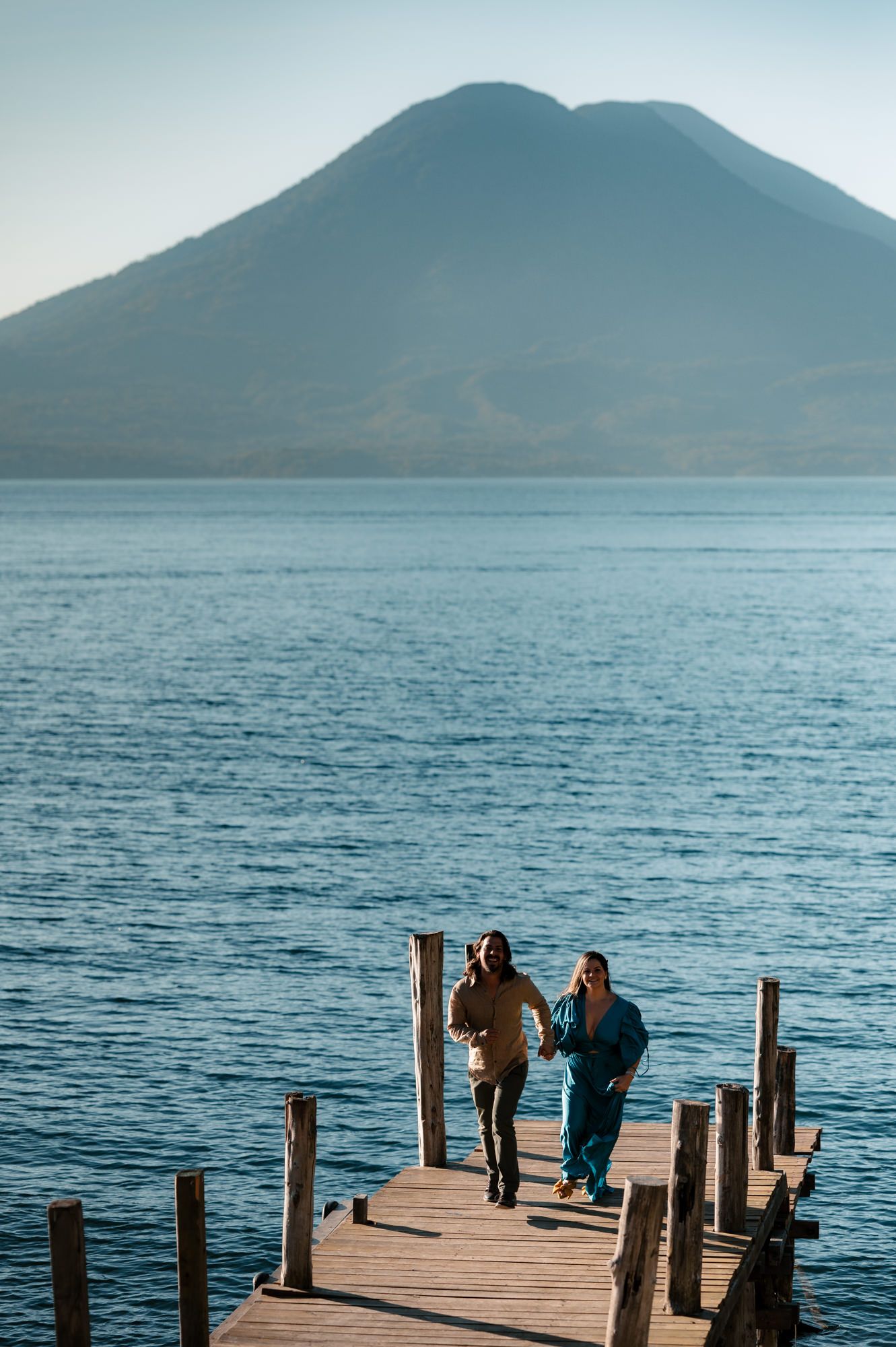 People stand on a wooden dock overlooking a serene mountain lake with blue waters and misty peaks in the distance.