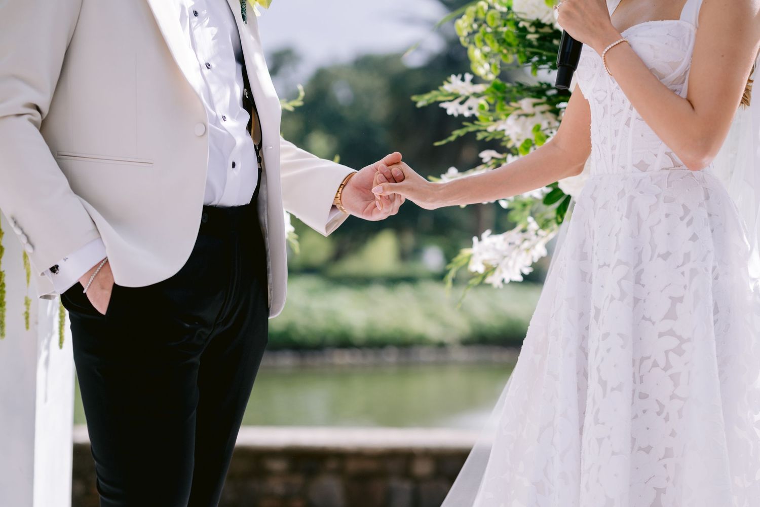 Close-up of couple holding hands during wedding ceremony with white lace dress detail.