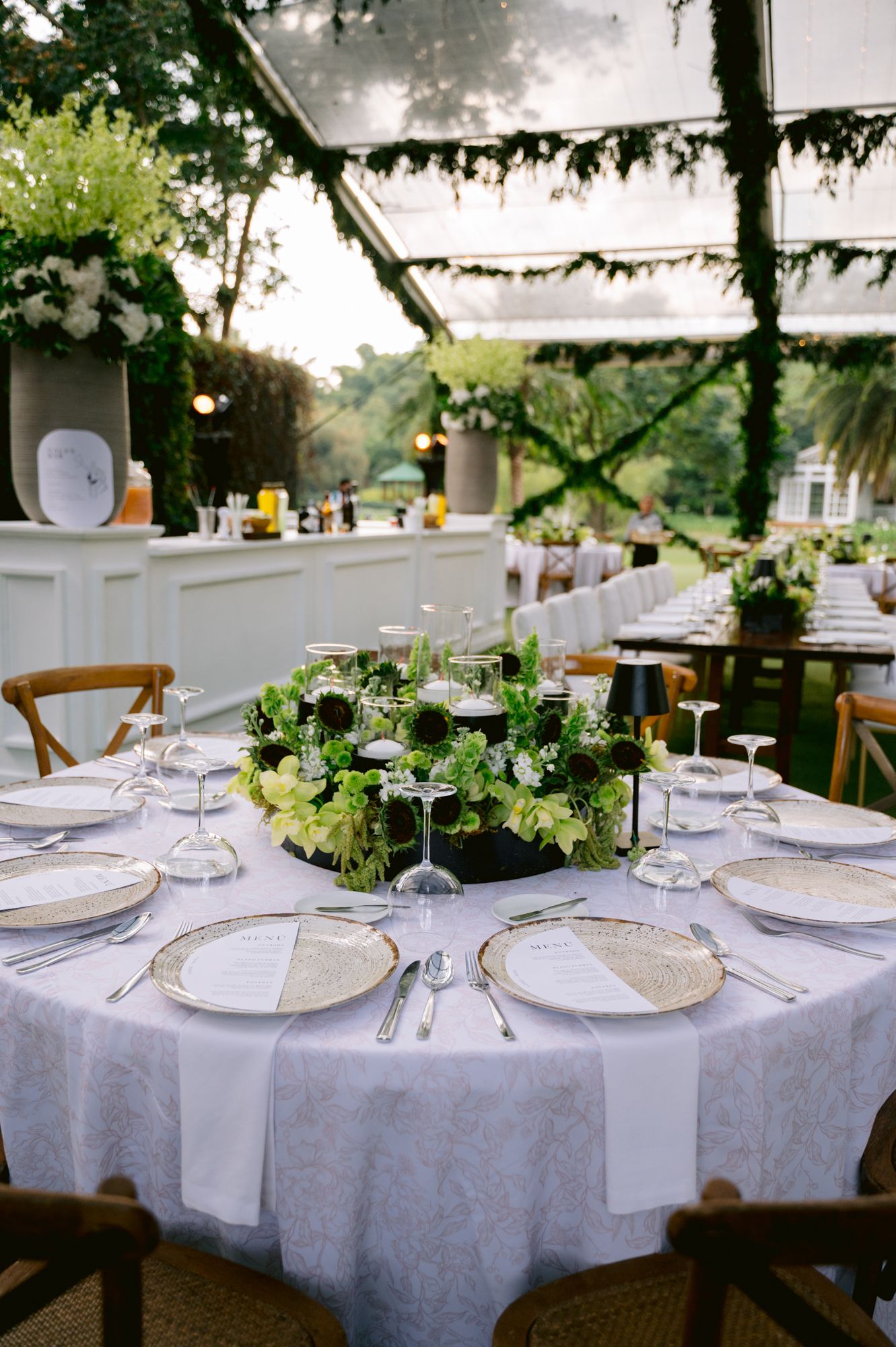 Elegant outdoor wedding reception tables set with white linens, glassware, and delicate floral centerpieces under hanging greenery.