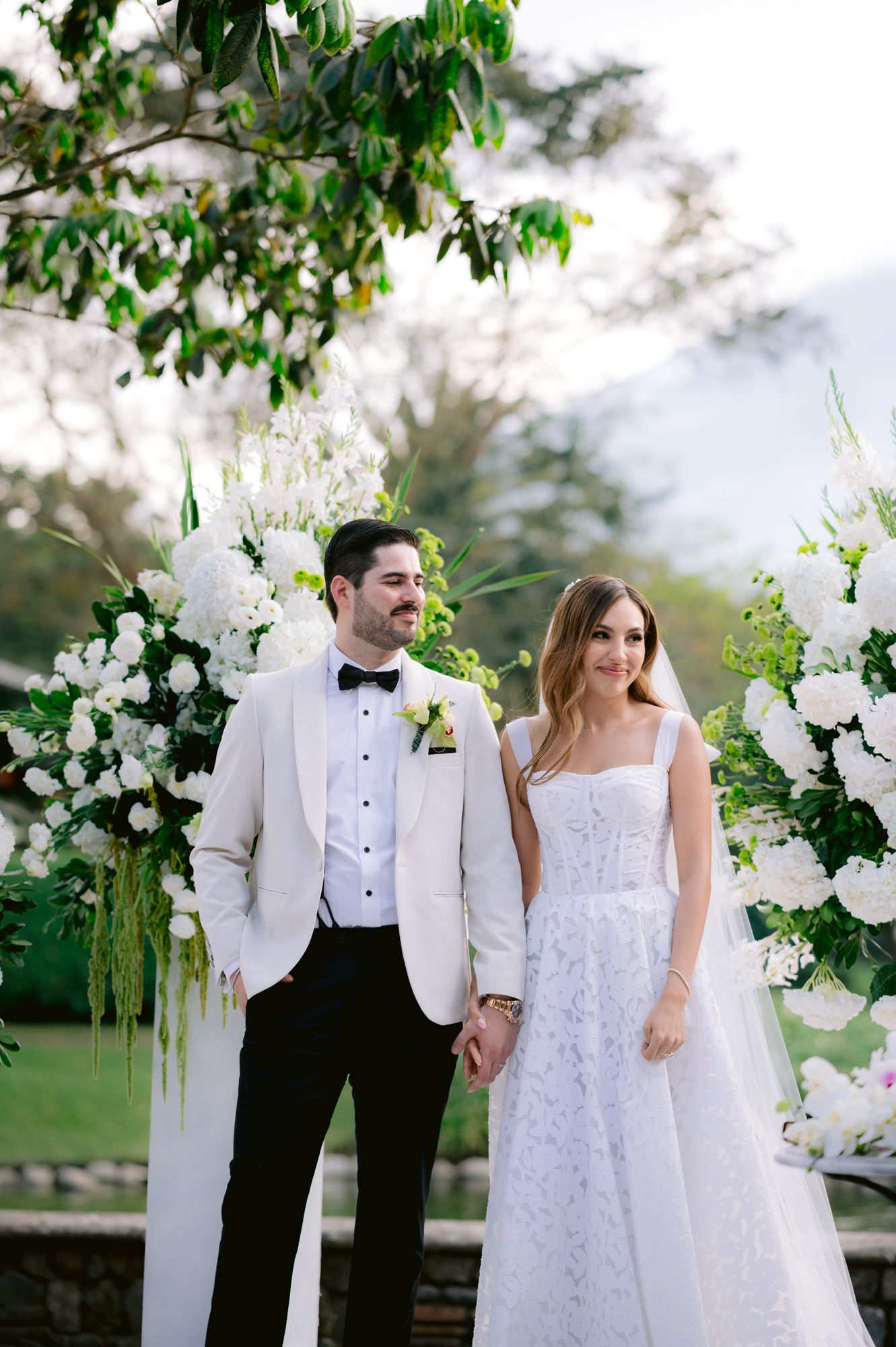 Newlyweds pose by white floral arch with groom in white jacket and bride in lace wedding dress.
