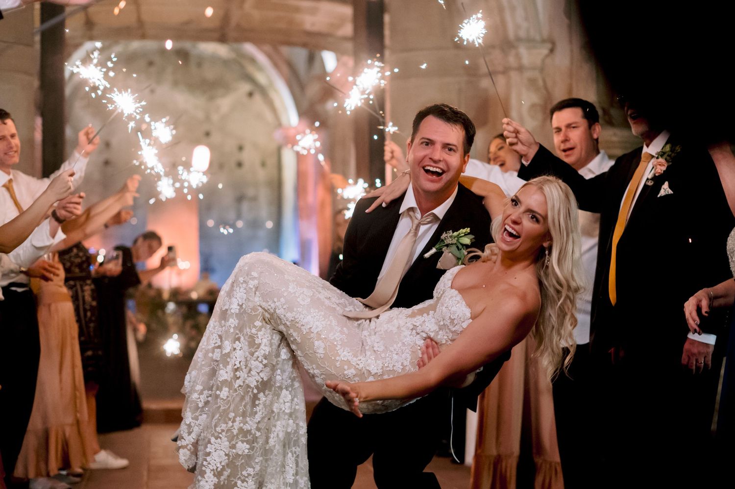 Joyful sparkler exit moment with groom carrying bride during wedding celebration.