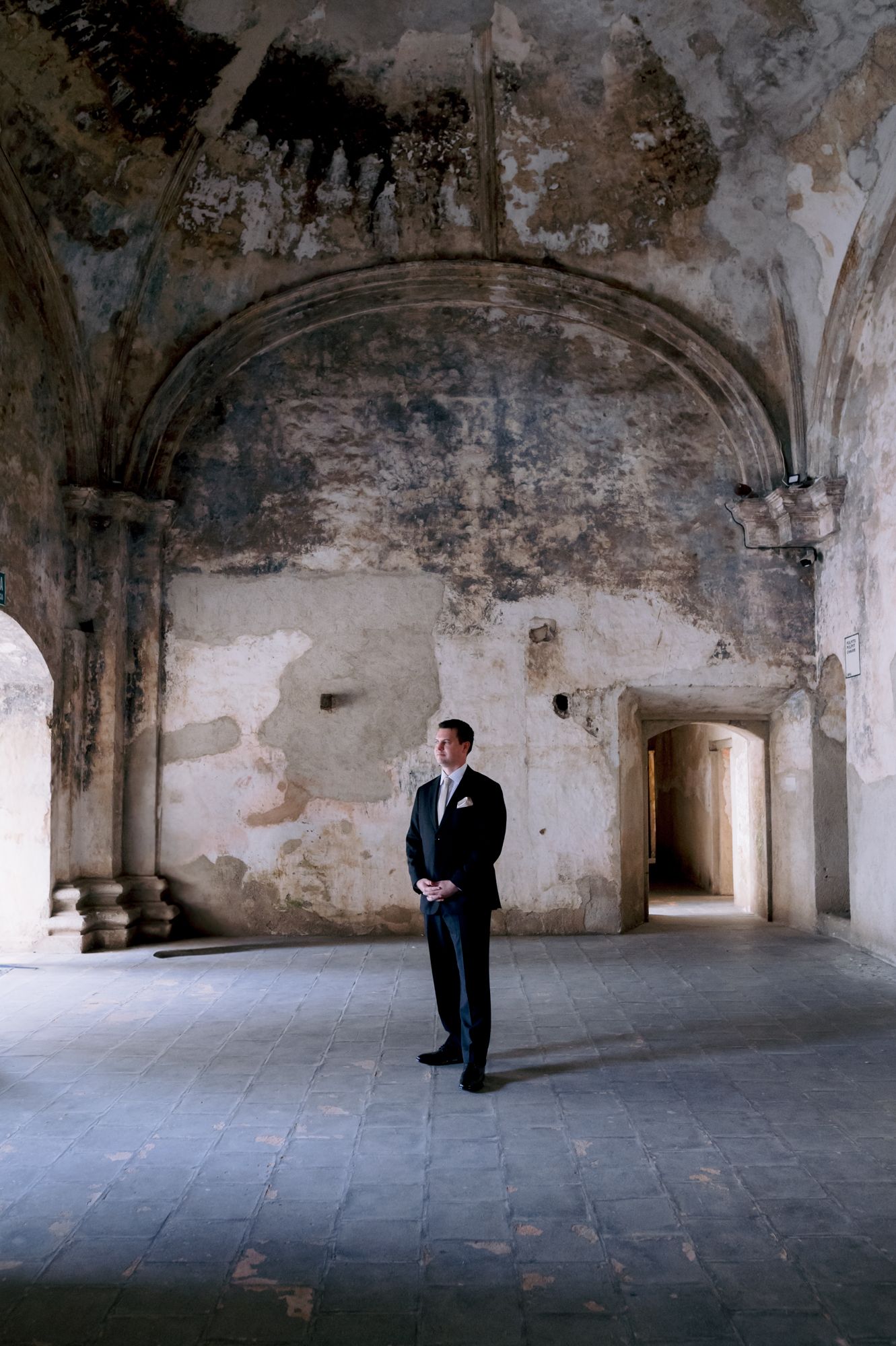Groom in classic black tuxedo stands in weathered stone hallway with arched ceiling.