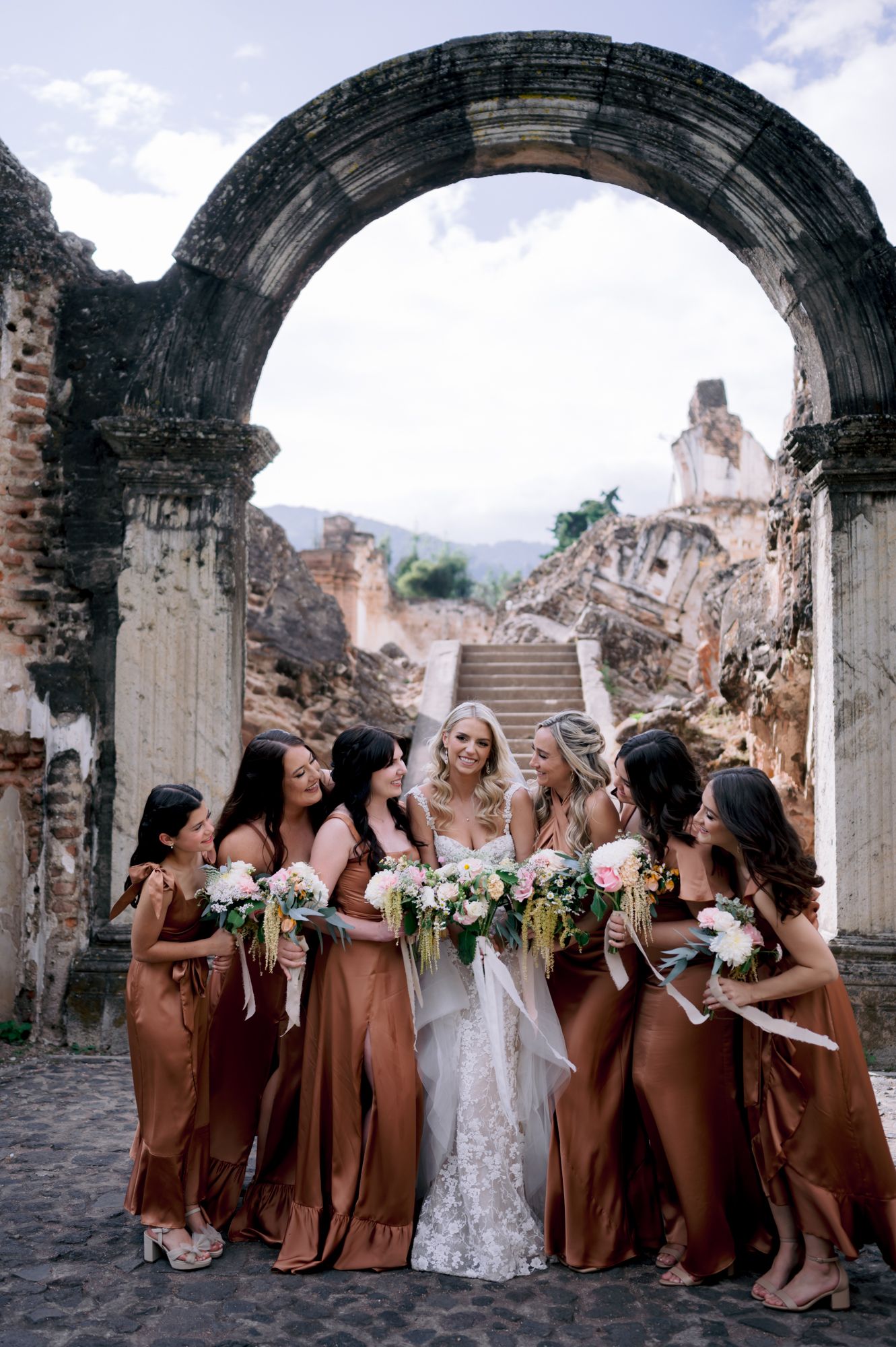 A bridal party in copper-colored dresses gathers under a stone archway at ancient ruins.