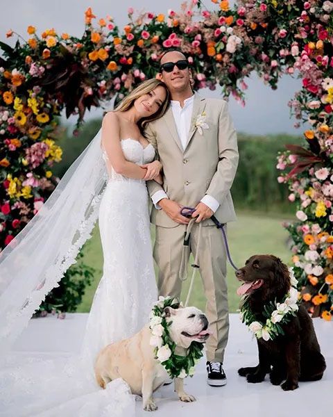 A outdoor wedding ceremony under a colorful floral arch with two adorned dogs joining the celebration.