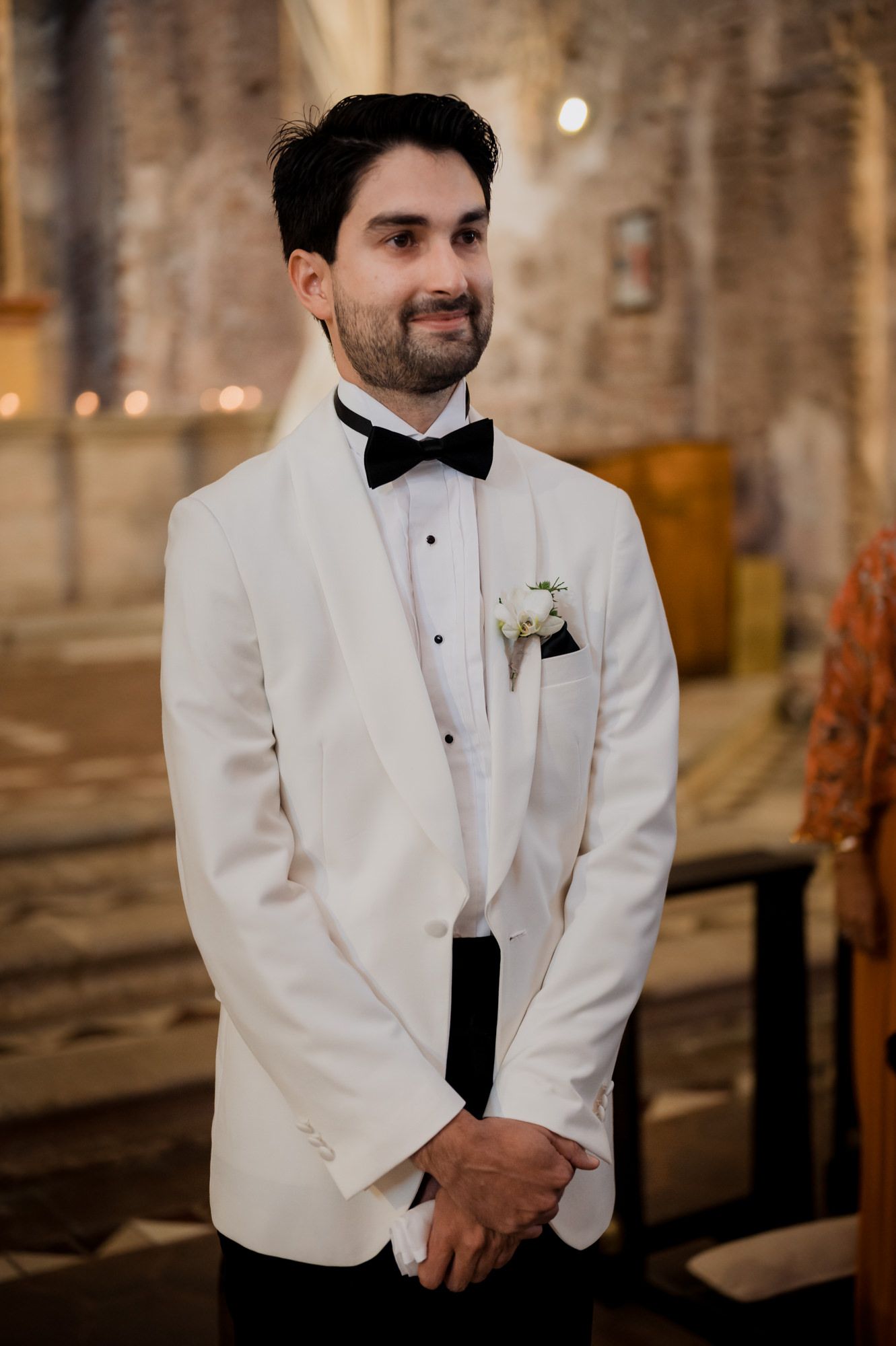 A groom in a white dinner jacket and black bow tie poses in a rustic stone venue.