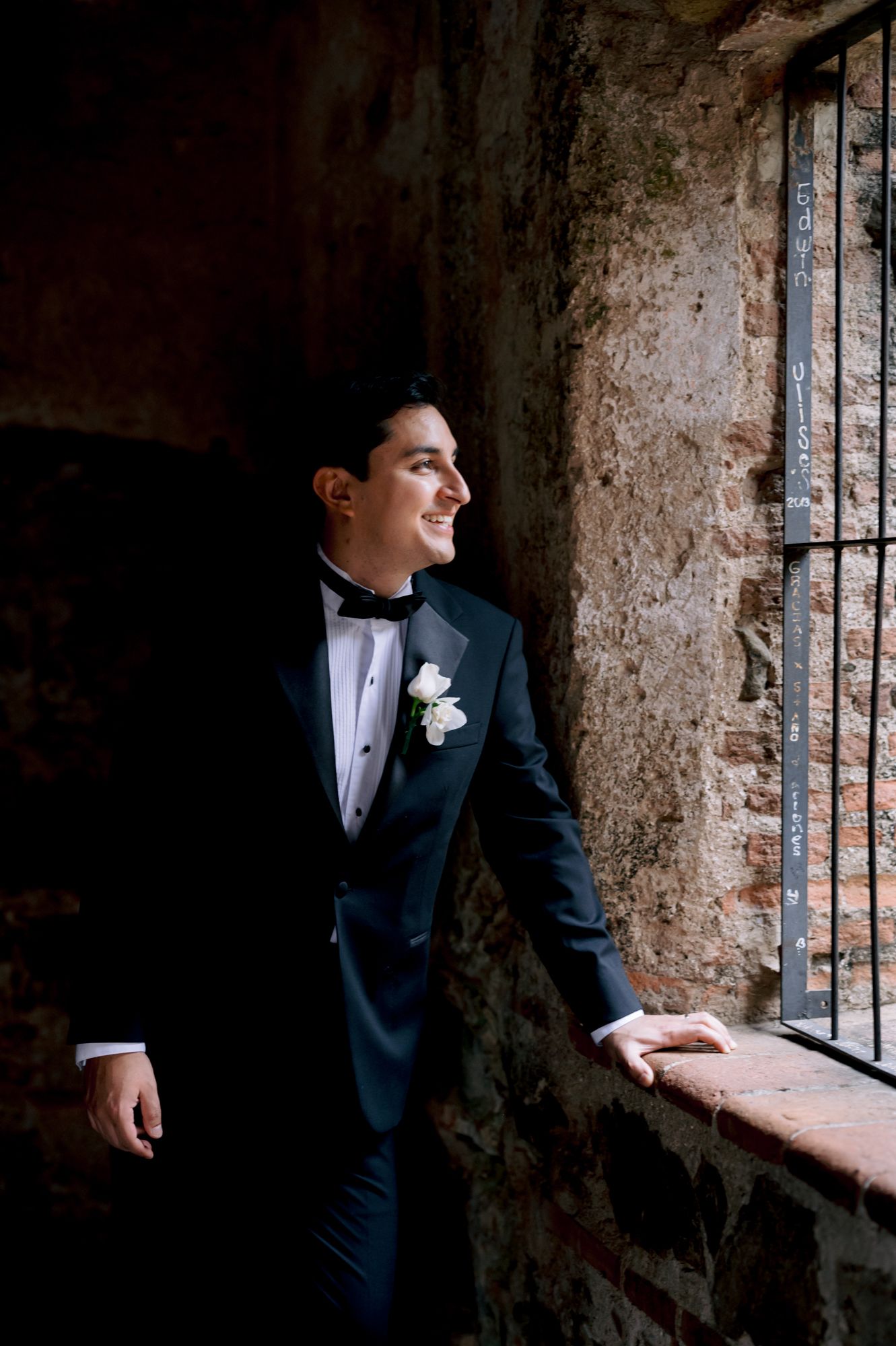 Groom in classic tuxedo poses by rustic window in historic brick building.