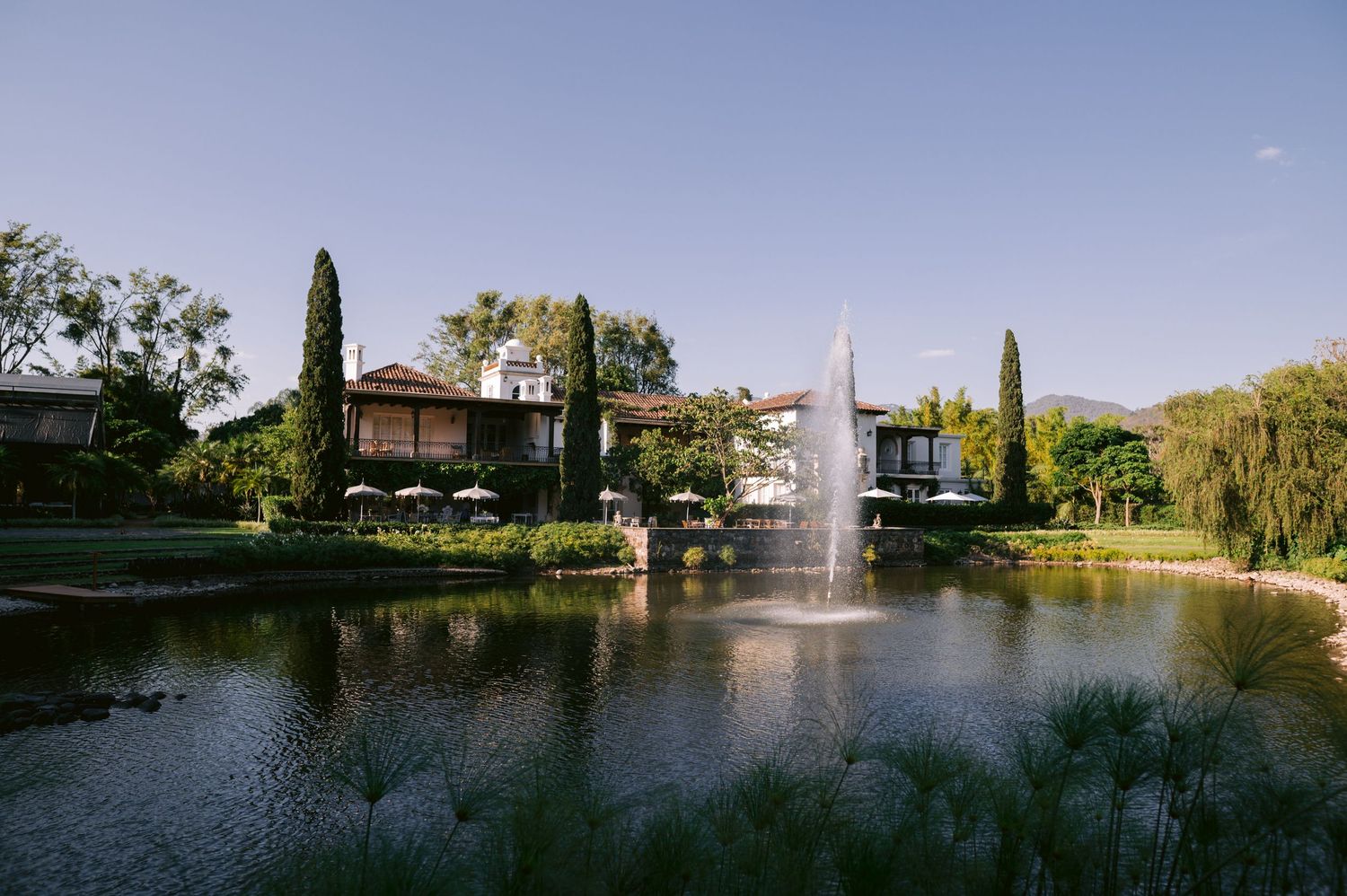 Serene garden landscape with a fountain in a pond surrounded by cypress trees and elegant architecture.